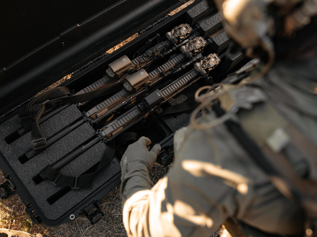 A close-up action shot of a person in tactical gear reaching into a black hard case containing several tactical rifles with mounted optics, set outdoors on dry grass.