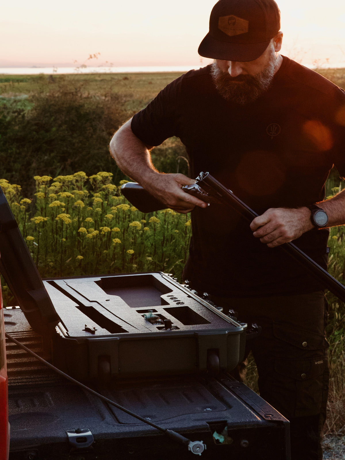 A bearded man in a black t-shirt and cap stands outdoors in a field at sunset, assembling a long gun while a black hard case sits open on a truck bed in the foreground.