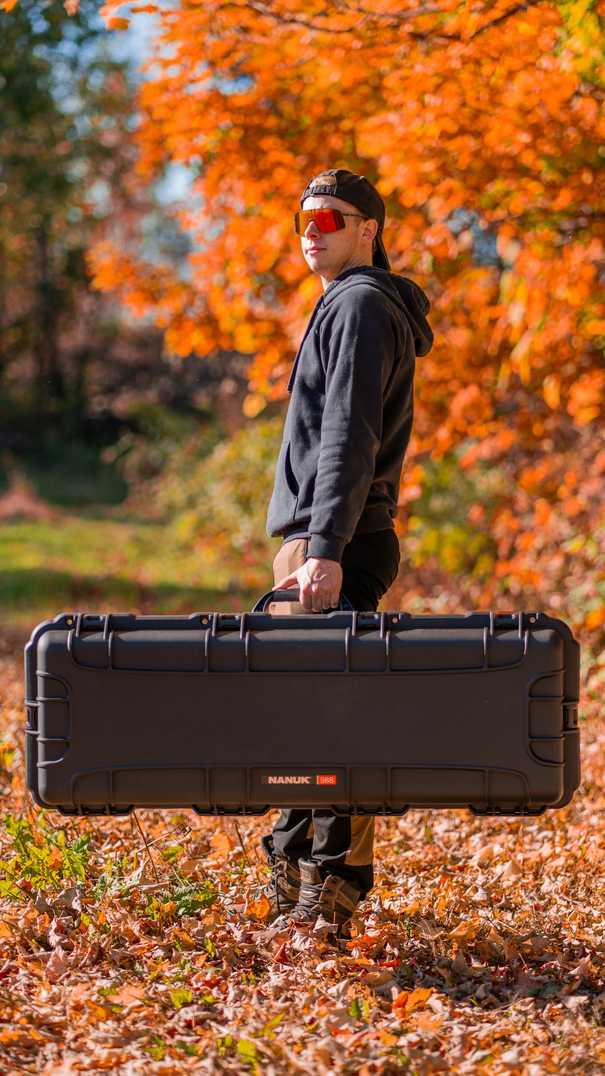 A man wearing a black hoodie, black cap, and orange-tinted sunglasses carries a large black hard case outdoors in an autumn setting with orange leaves on the ground and trees in the background. The brand 'MANUKE' is visible on the front of the case.