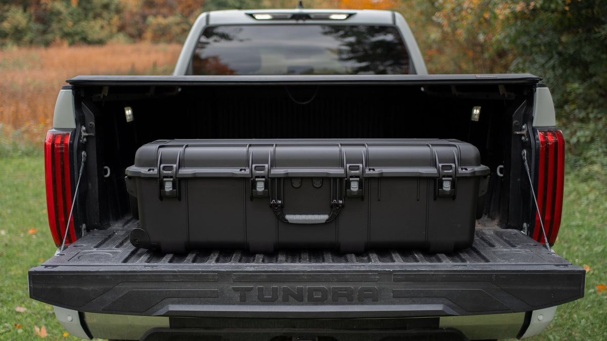 A large black hard-sided storage case loaded into the bed of a silver pickup truck, shown from the rear with the tailgate down. The truck is parked outdoors on grass with trees in the background.