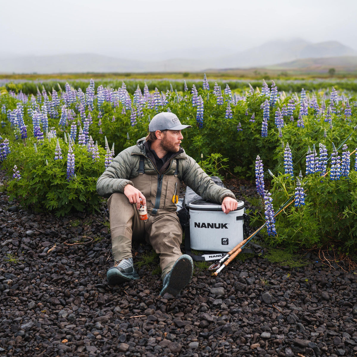 A man in outdoor fishing attire sits on dark gravel beside a white NANUK cooler, with a fishing rod and a field of purple lupines in the background.