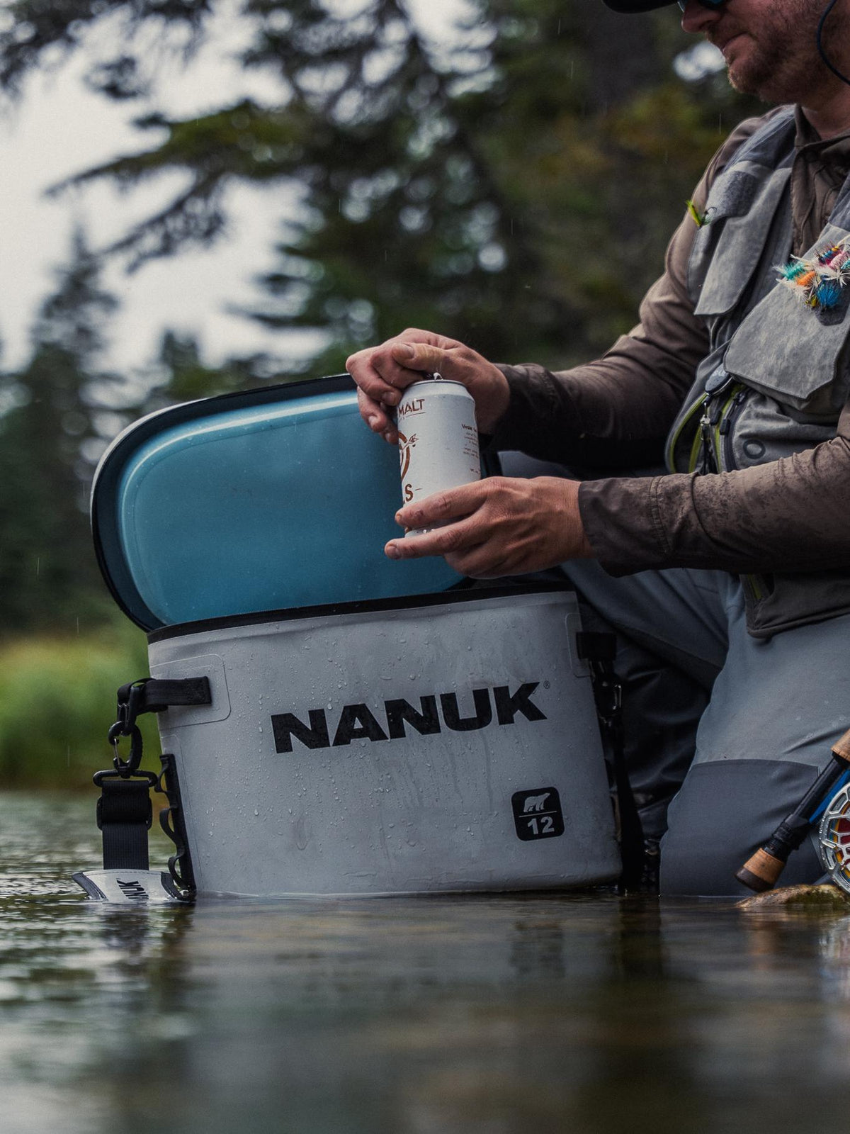 A person in a fishing vest sits in shallow water beside a large gray NANUK cooler with a blue lid, holding a white can.