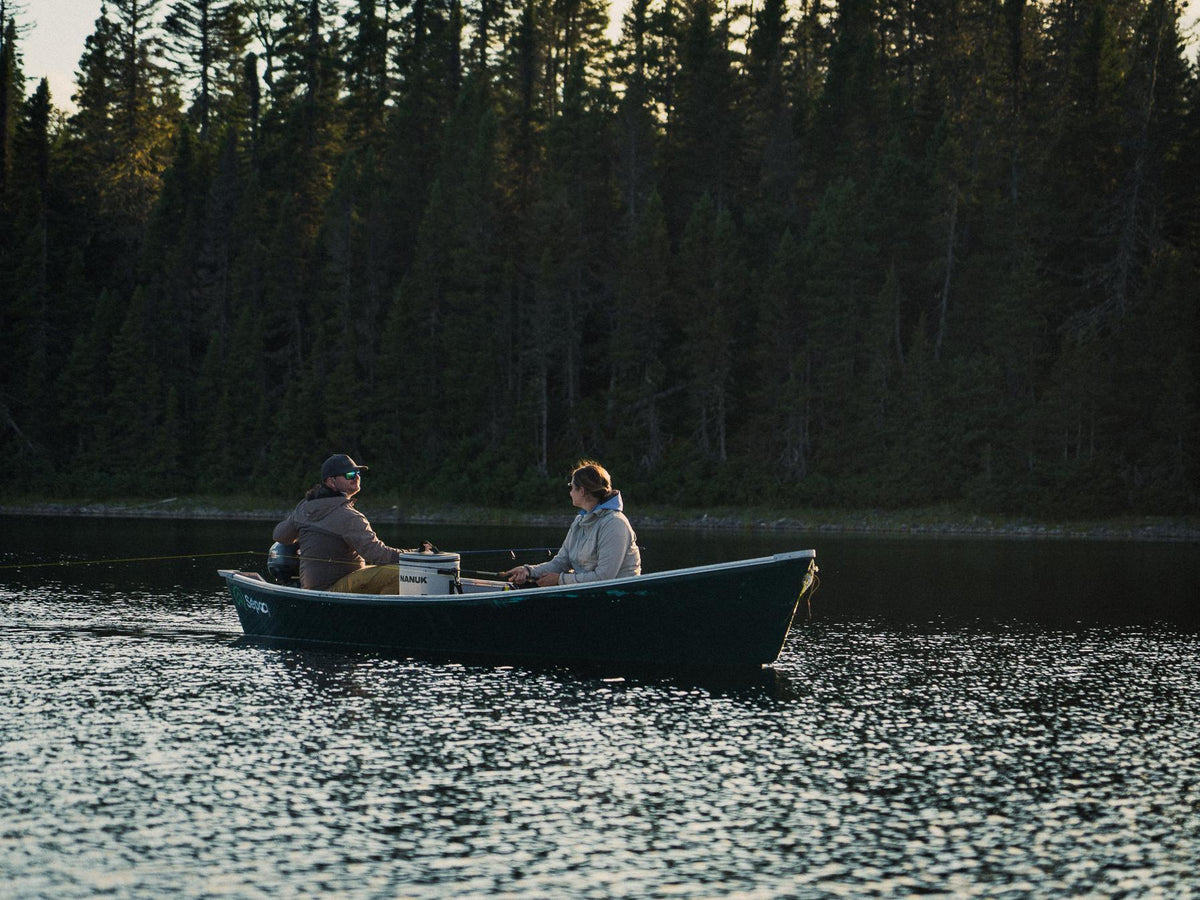 Two people sitting in a small rowboat on a lake with a dense evergreen forest in the background at sunset.