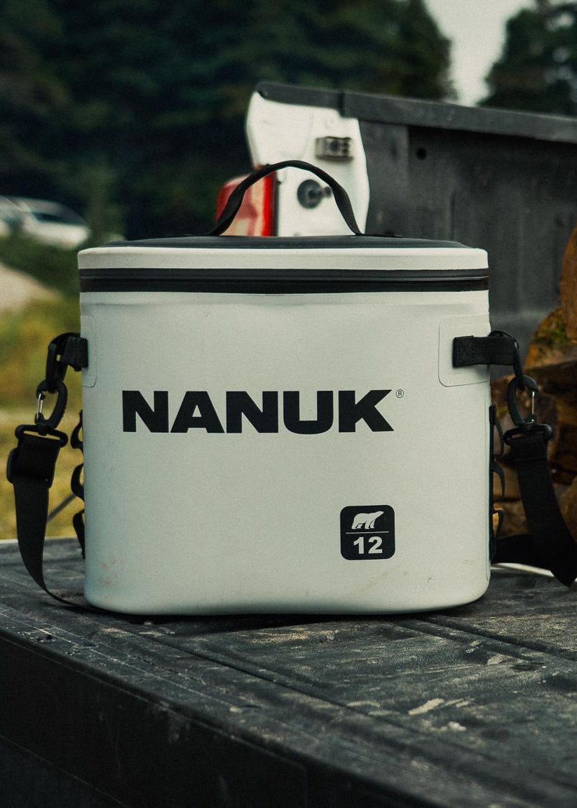 A white Nanuk 12-liter cooler with black straps and branding sits on the bed of a truck next to a stack of firewood.
