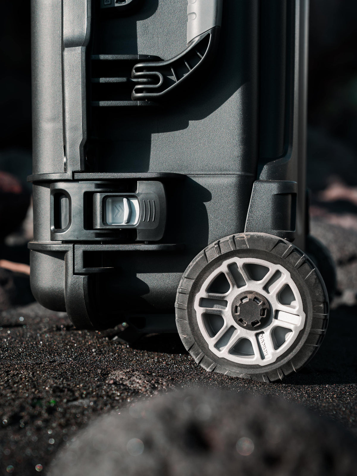 Close-up of a rugged black hard-shell suitcase with a large spoked wheel and textured treaded tire, standing on dark ground in dramatic outdoor lighting.