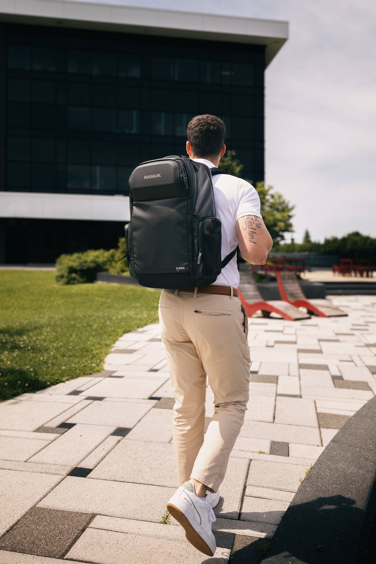 Man walking outdoors with a black backpack, wearing a white shirt and beige pants.
