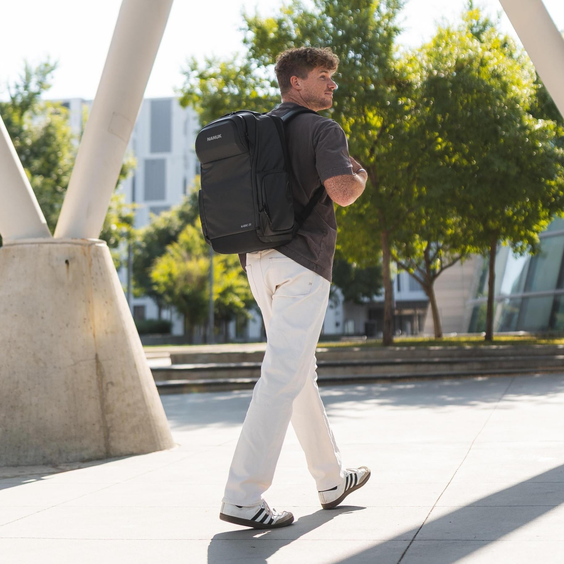 Man walking outdoors with a backpack, surrounded by trees and modern architecture.