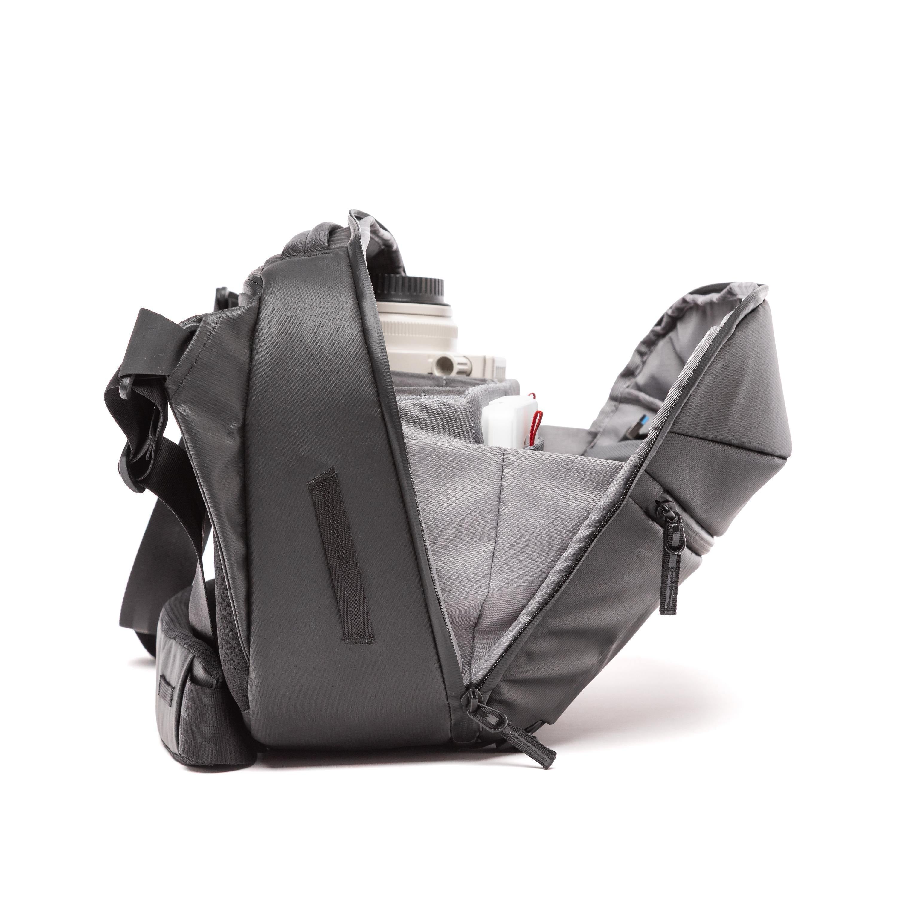 A dark gray camera shoulder bag shown open, displaying its interior compartments and a camera lens inside, set against a plain white background. A dark gray camera shoulder bag shown open, displaying its interior compartments and a camera lens inside, set against a plain white background.