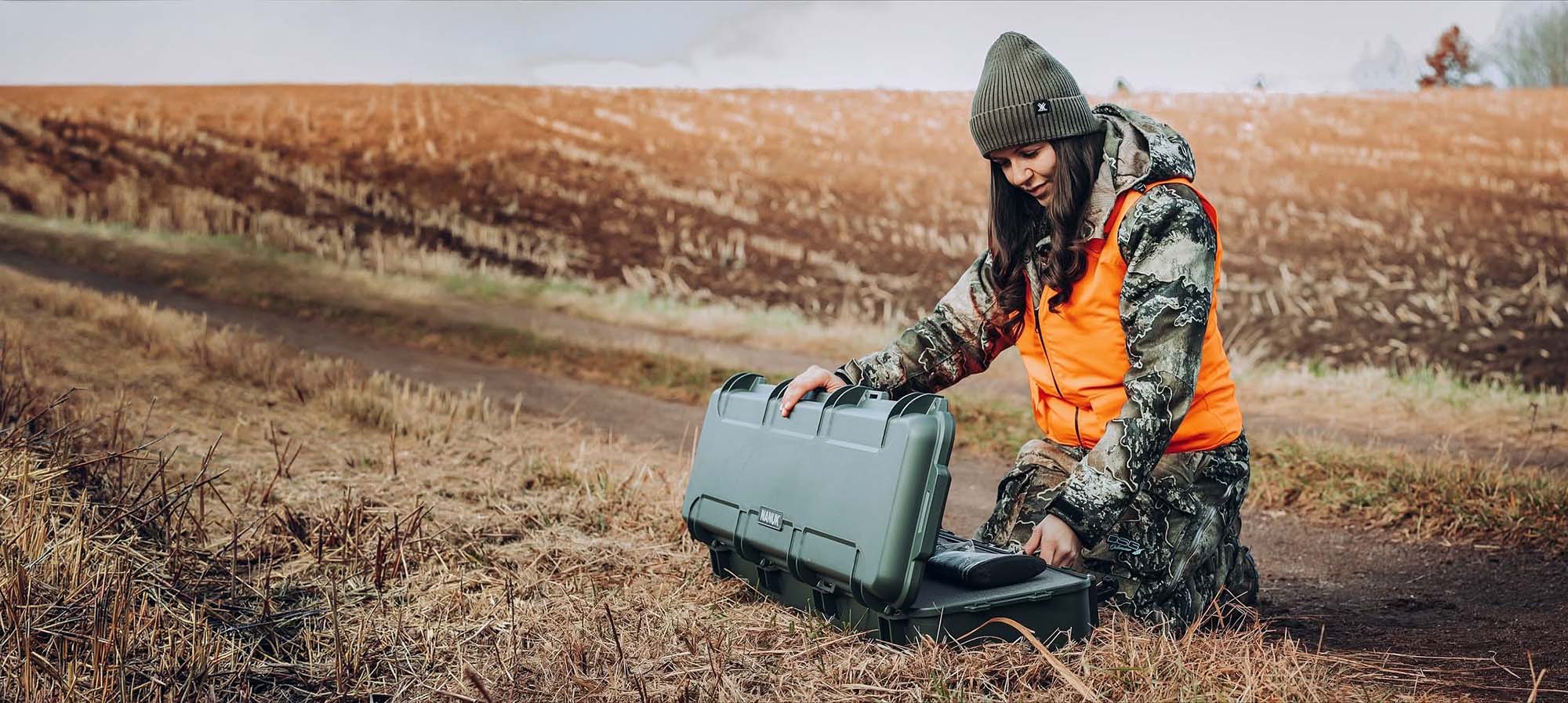 A woman in hunting attire kneels in a field, opening a protective Nanuk case filled with gear, perfect for nanuk cases hunting and outdoor promotion.