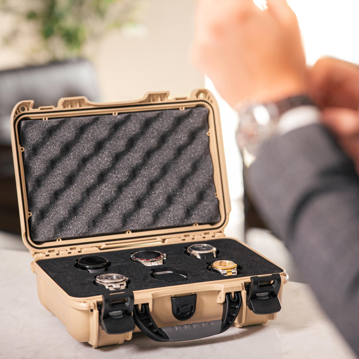 A tan hard case with black foam interior holding several wristwatches, shown open in a real indoor setting with a person's arm blurred in the background.