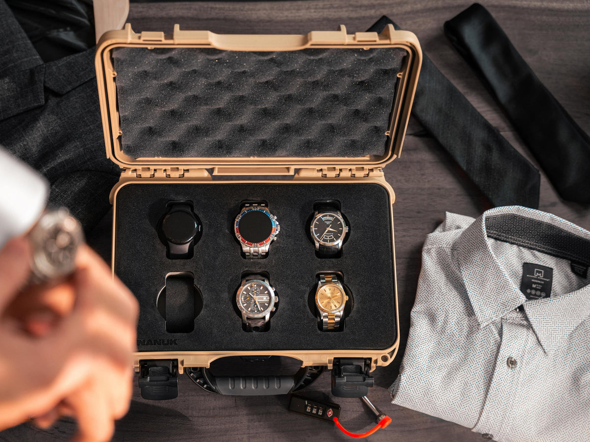 A tan hard case with black foam insert holding six wristwatches, styled on a dark tabletop with a folded light shirt, a black tie, and a hand holding a watch in the foreground.