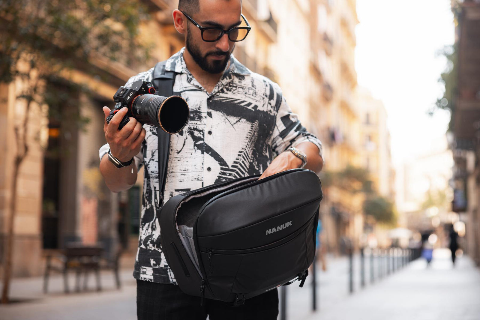 A man wearing a black and white patterned shirt and glasses walks down a city street while holding a camera and looking into the pocket of a black HANUK backpack.