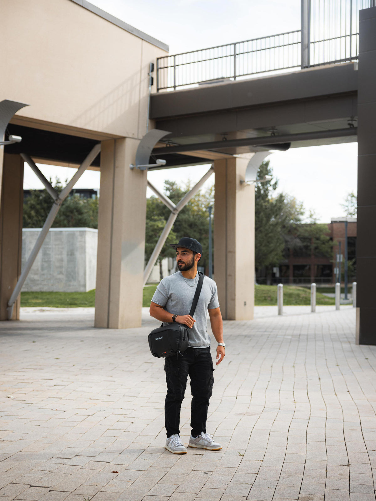 A man wearing a gray t-shirt, black pants, and a black cap stands outdoors under a modern concrete walkway, carrying a black shoulder bag.