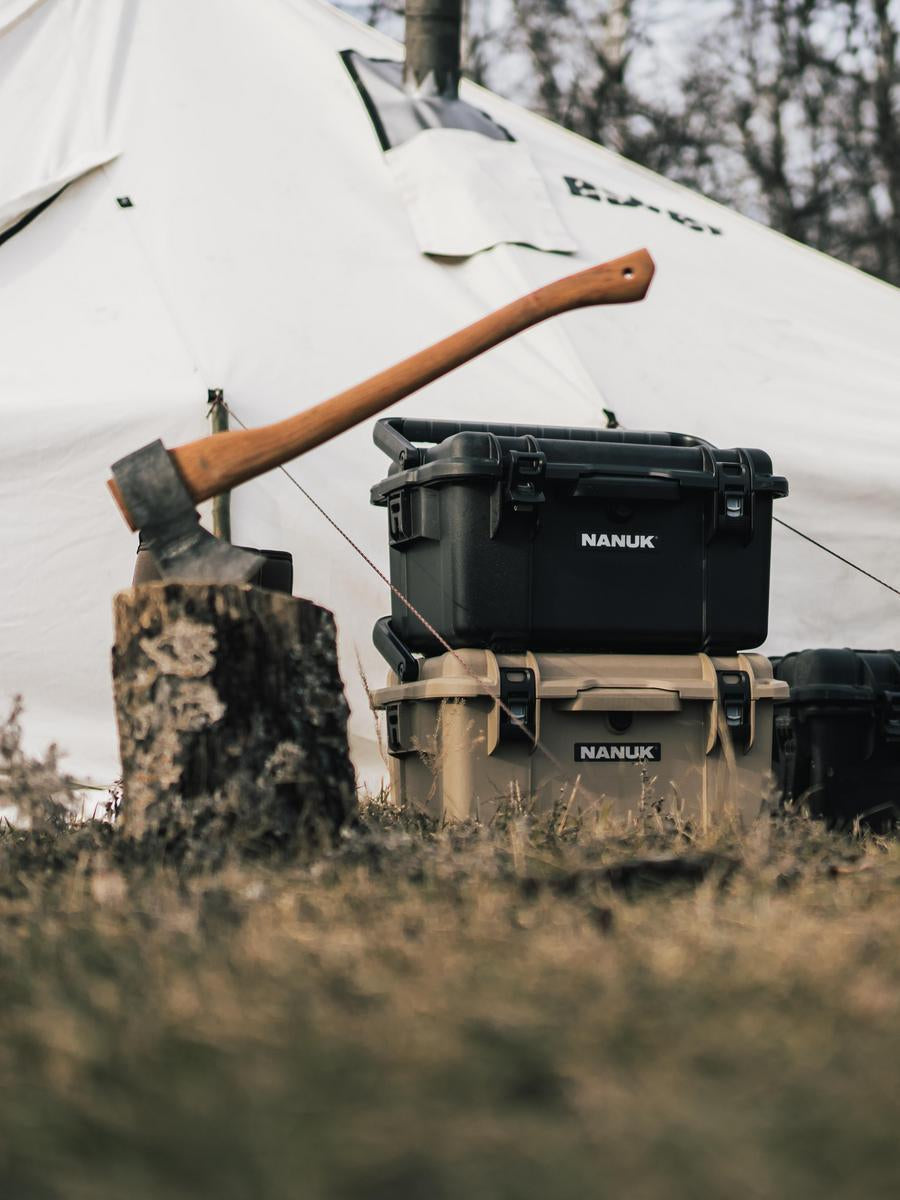 Black Nanuk hard-shell backpack with an axe on a stump in front of a white tent.