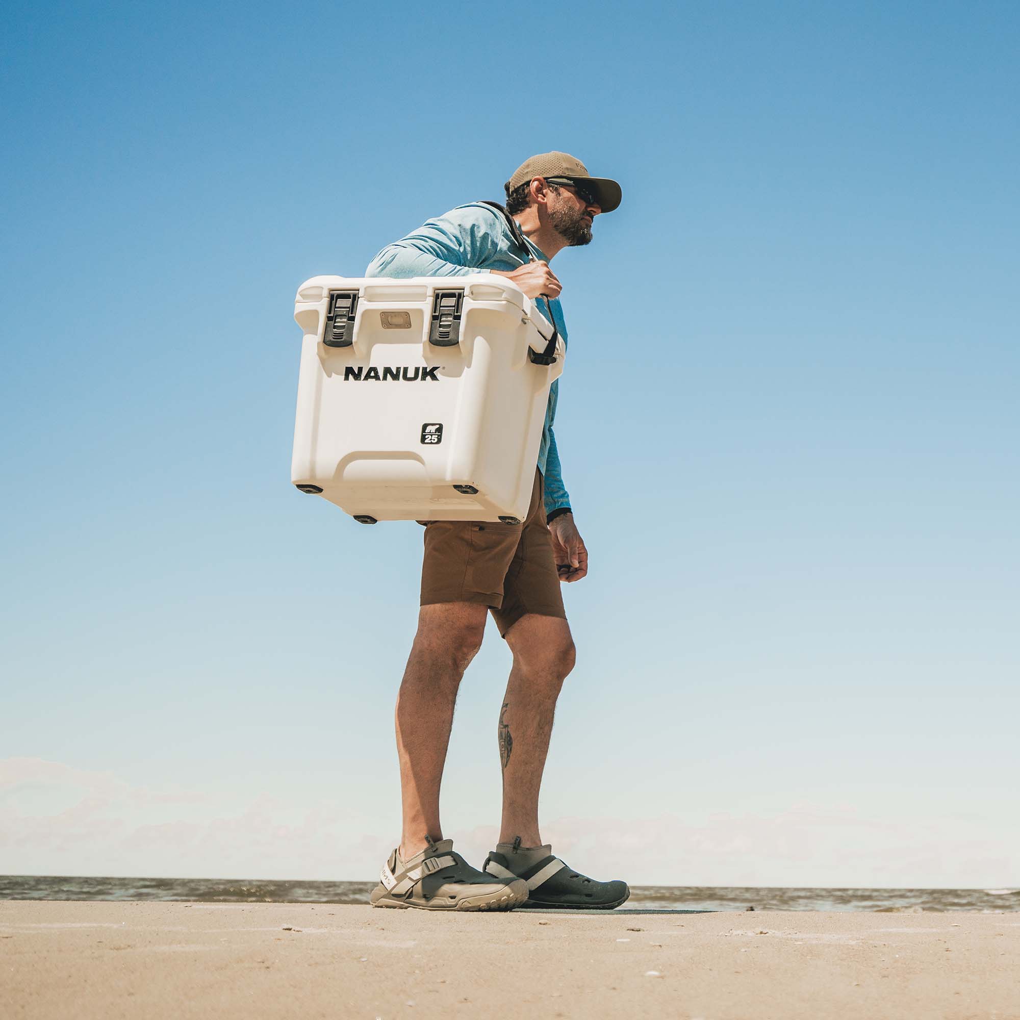 A man carrying a large white NANUK cooler on his shoulder, walking outdoors against a clear blue sky. A man carrying a large white NANUK cooler on his shoulder, walking outdoors against a clear blue sky.