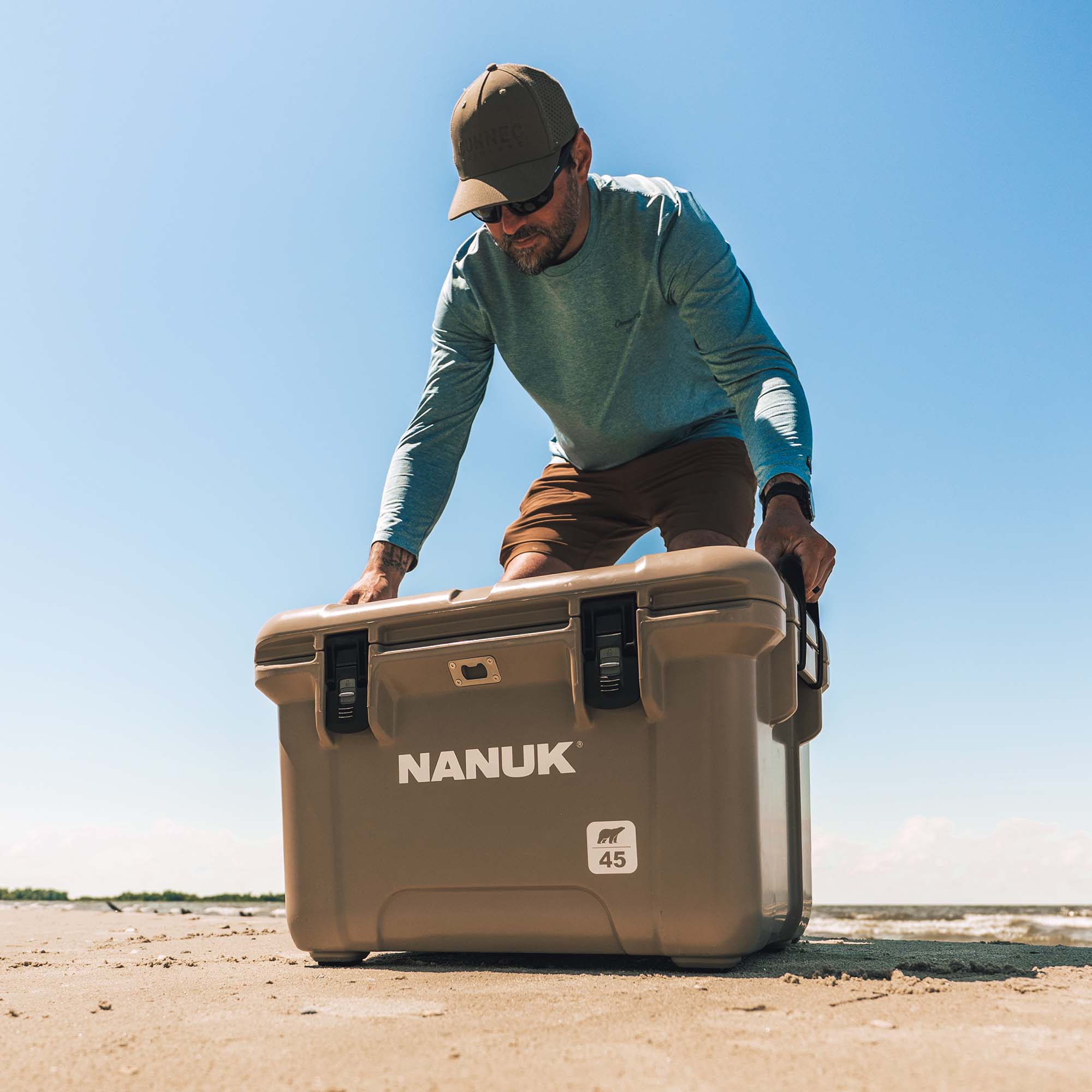 A man wearing a cap and sunglasses pushes a large tan Nanuk cooler on a sandy beach under a clear blue sky.