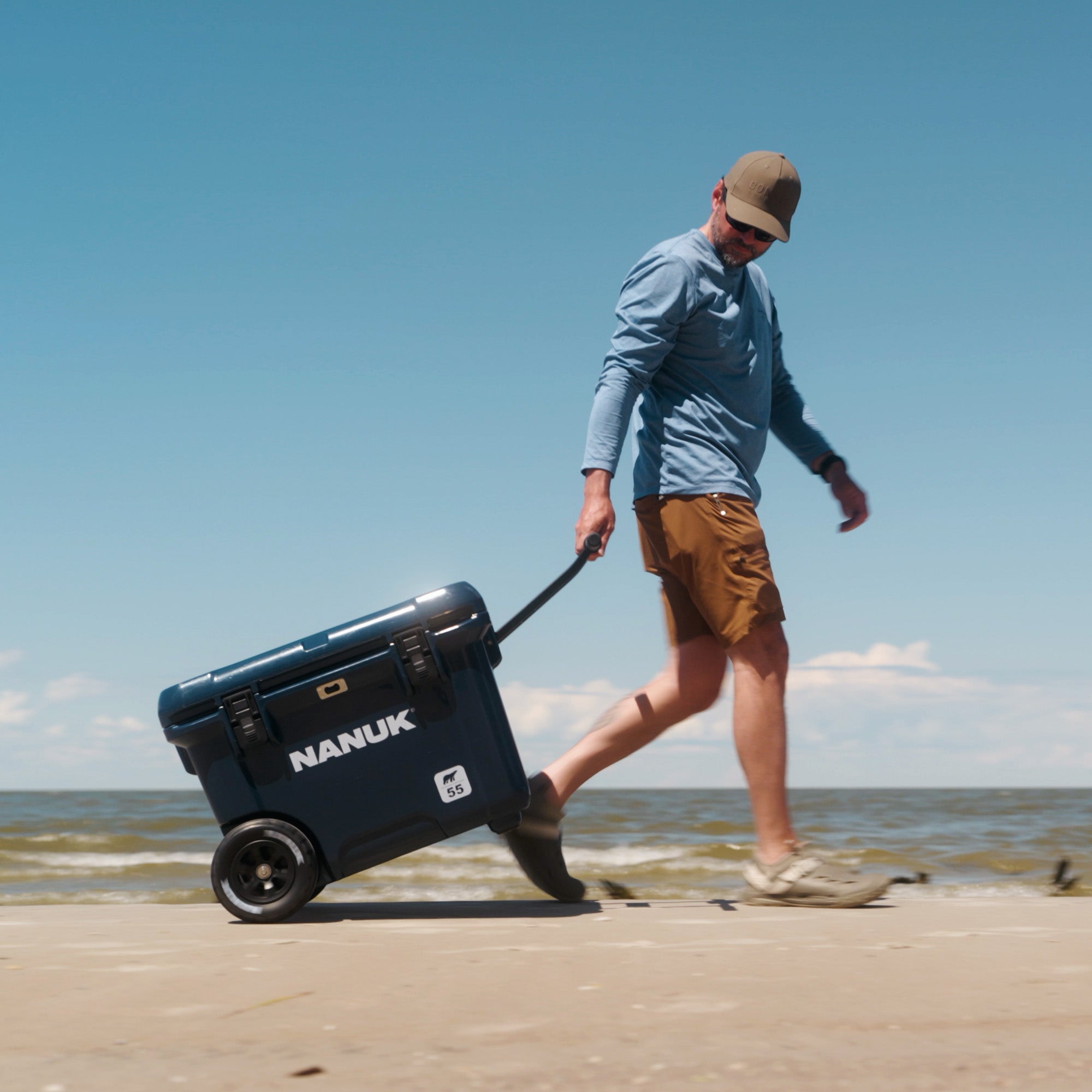 A man walks along a sandy beach pulling a dark blue Nanuk cooler on a wheeled dolly under a clear blue sky. A man walks along a sandy beach pulling a dark blue Nanuk cooler on a wheeled dolly under a clear blue sky.