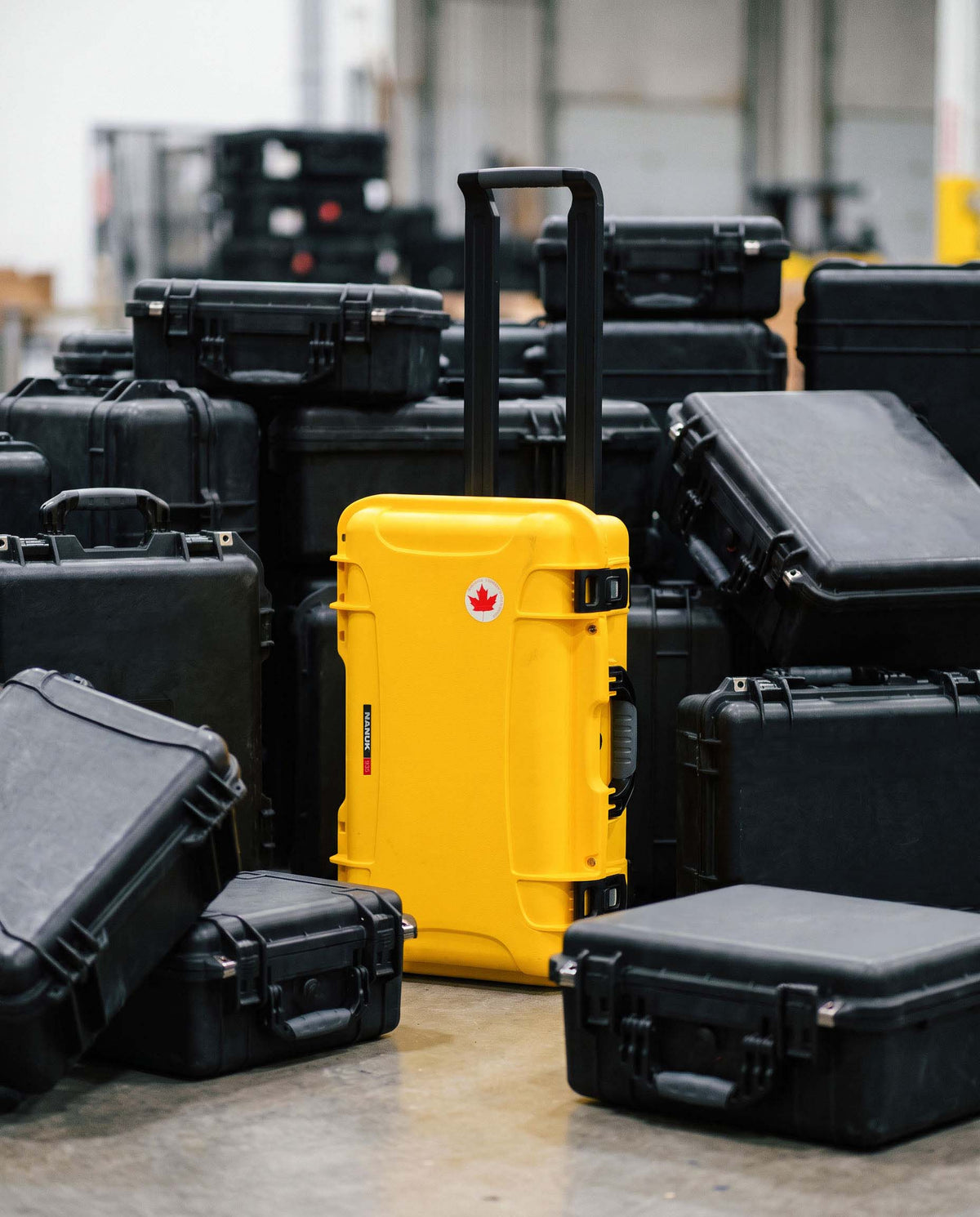 A bright yellow hard-shell rolling suitcase with an extended black handle stands in the center, surrounded by many black hard-shell cases in a warehouse-like setting.