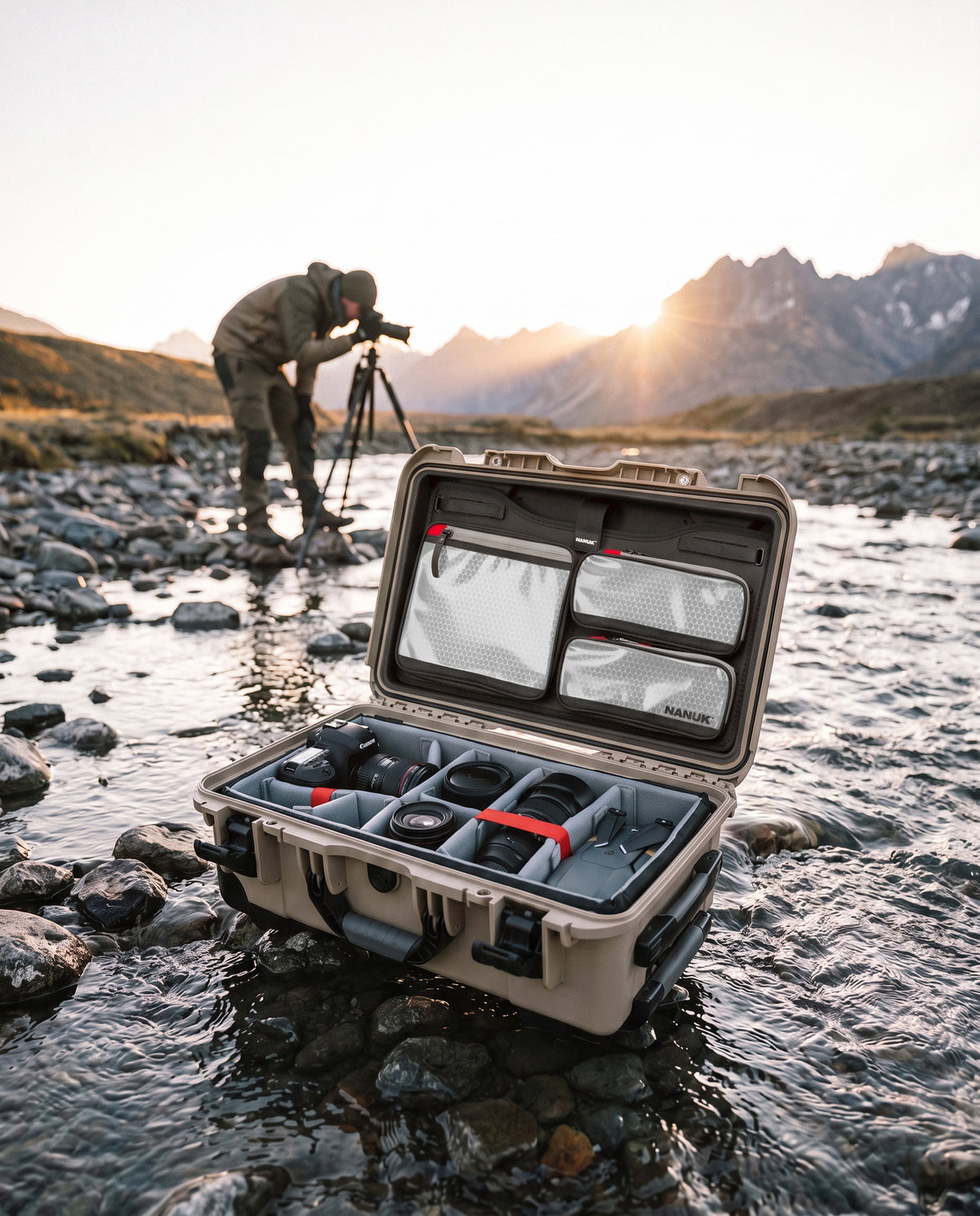 An open hard-shell camera gear case sits in shallow water on a rocky riverbank, with a photographer working in the background near a tripod and mountains at sunset.