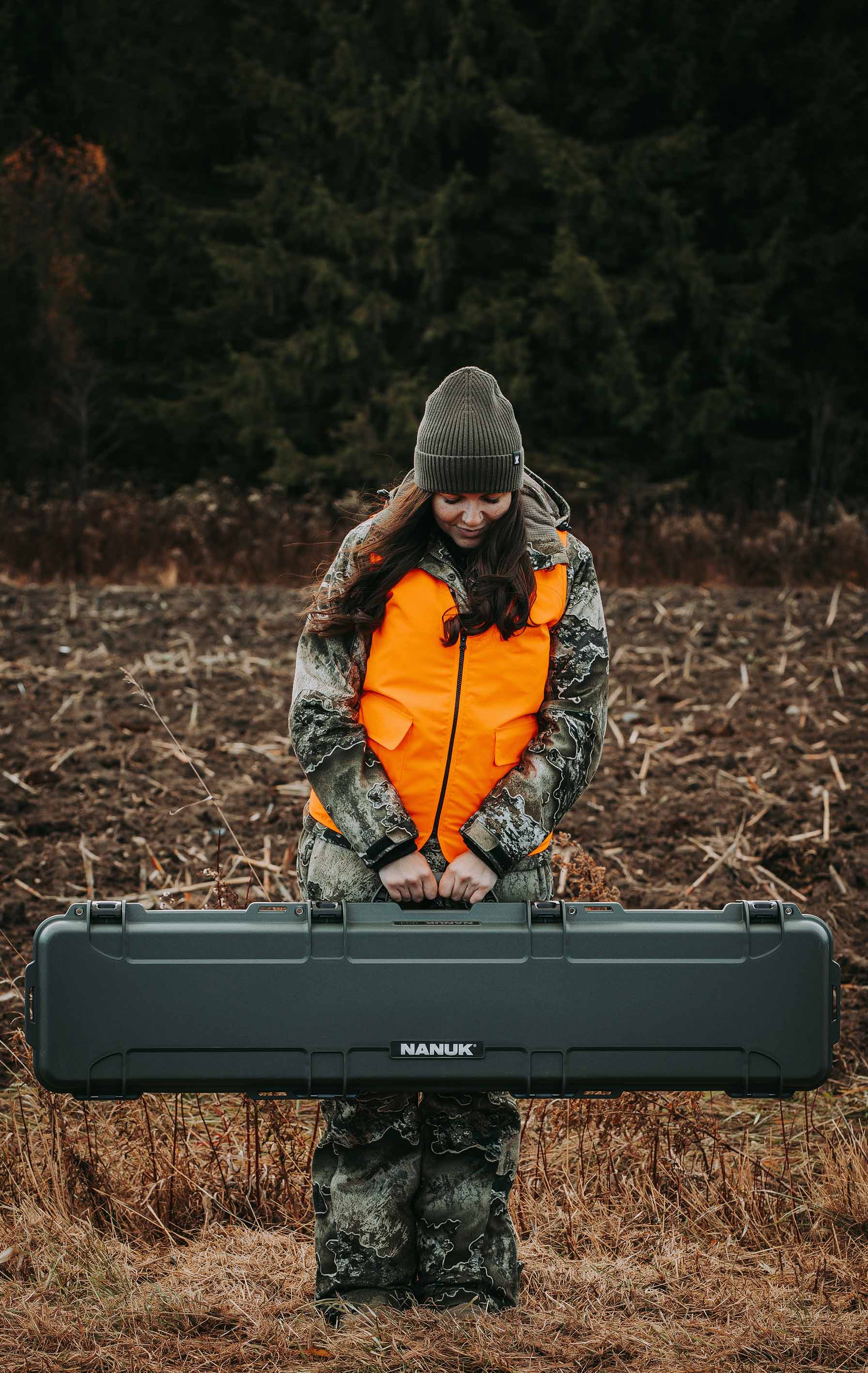 A smiling woman in hunting camouflage and an orange vest holds a Nanuk case outdoors, suitable for Nanuk cases hunting and outdoor promotion.
