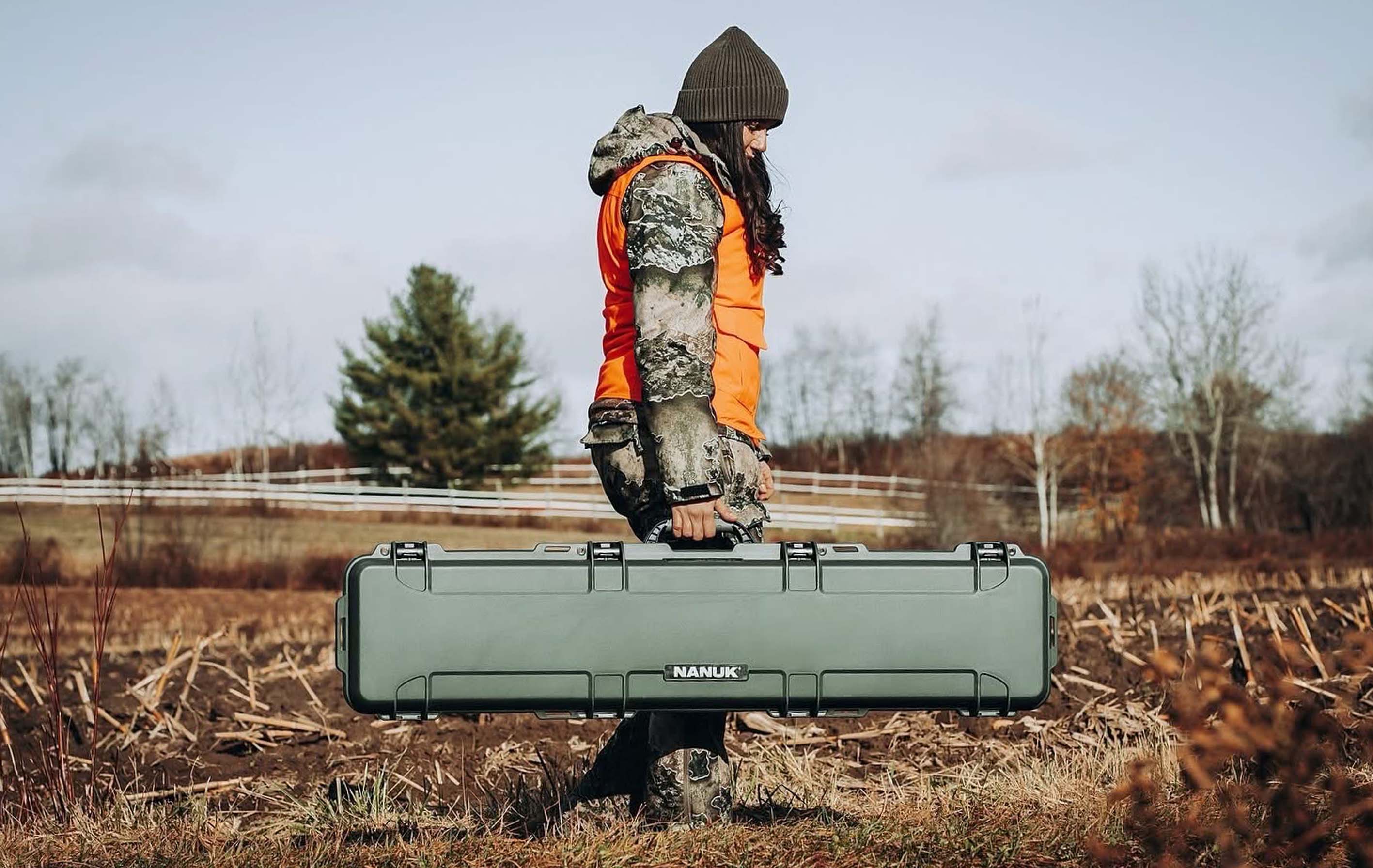 A person in camouflage and an orange vest carries an olive green Nanuk case through a field, serving as a nanuk cases hunting and outdoor promotion.