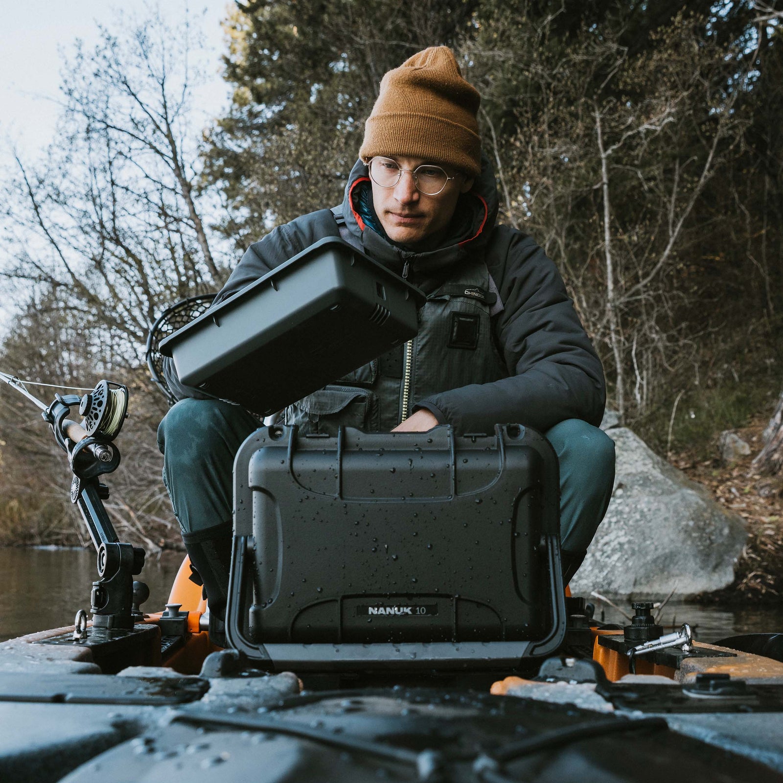 Angler in winter clothing kneeling in a kayak on a lake, holding a closed NANUK Trail 10 case.