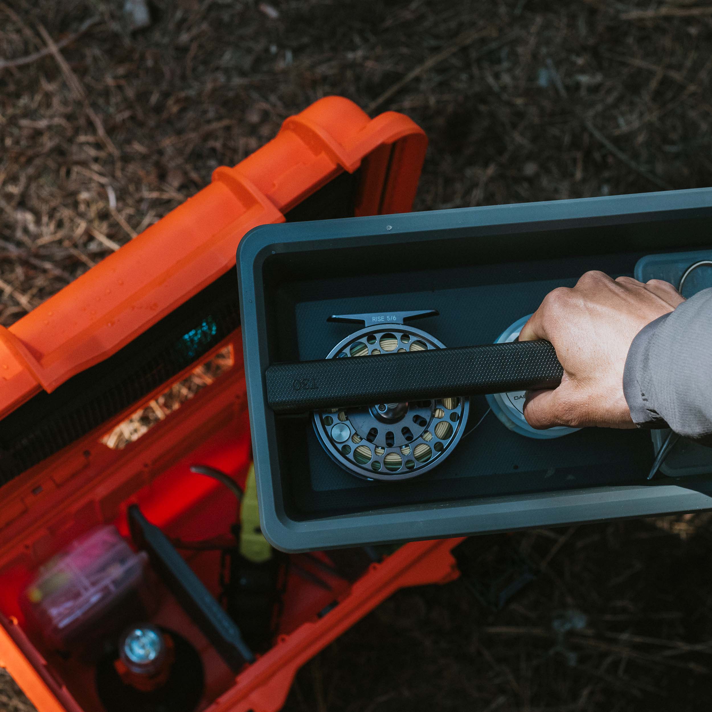 Interior shot of an open orange NANUK Trail 15 case on the forest floor, foam tray removed and a hand placing gear into the compartments. Interior shot of an open orange NANUK Trail 15 case on the forest floor, foam tray removed and a hand placing gear into the compartments.
