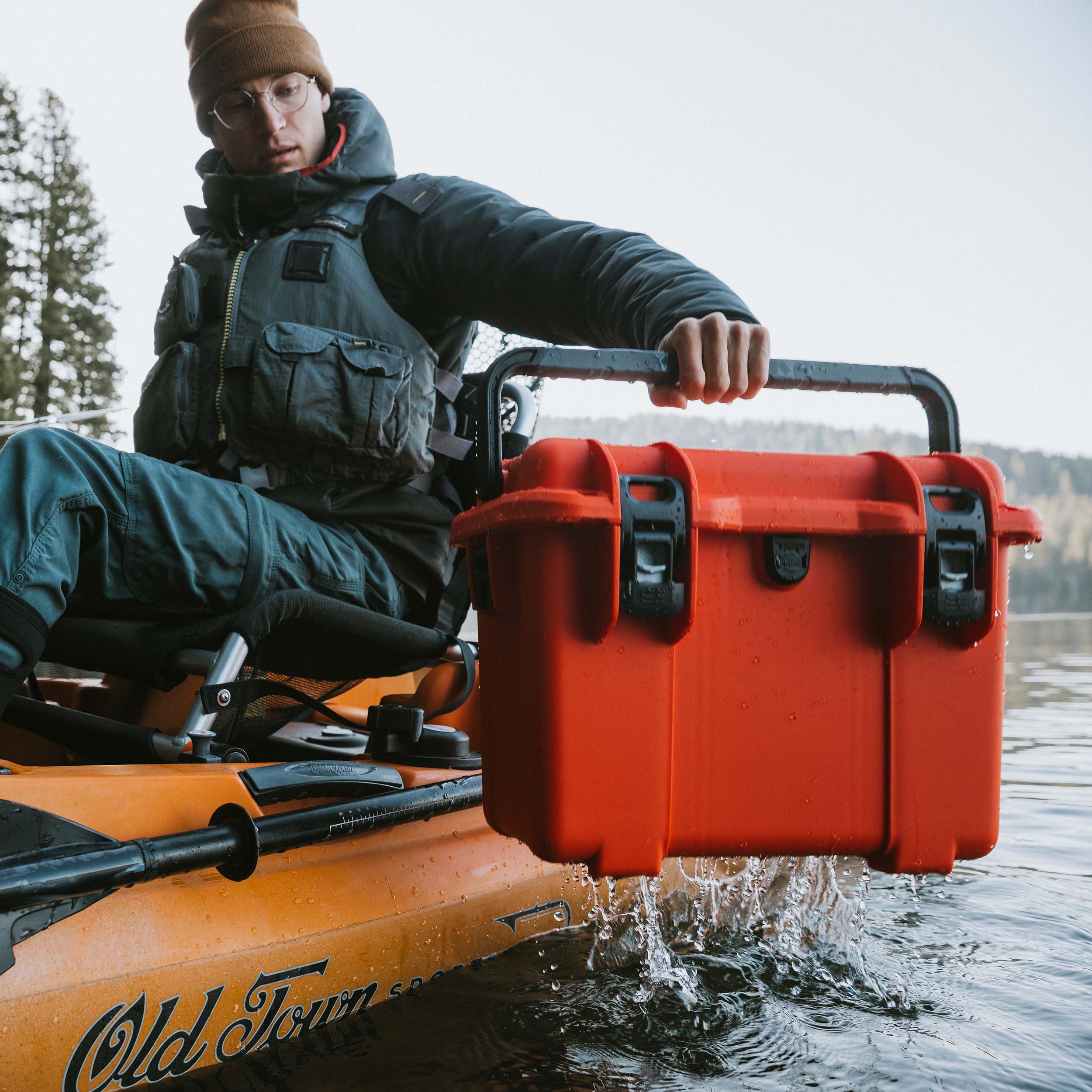 Person fishing from a kayak on a lake with a closed orange NANUK Trail 15 protective case beside them.