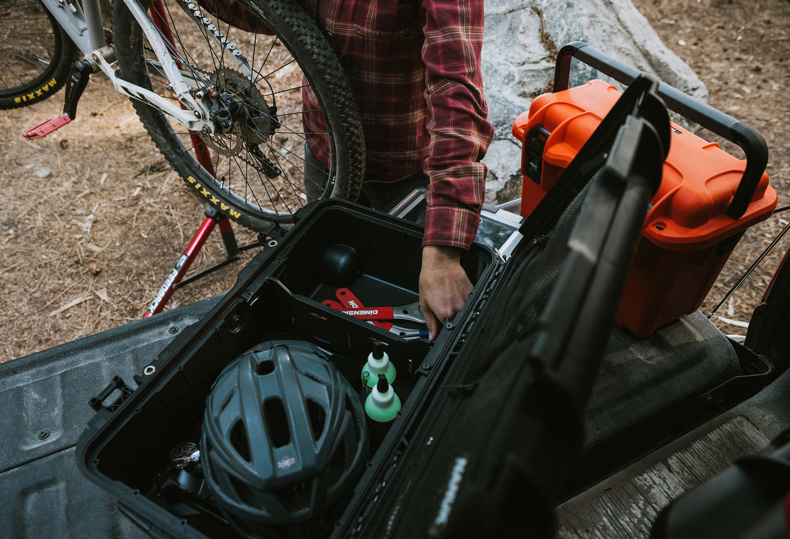Person loading a black NANUK Trail 30 case into the bed of a pickup truck next to a mountain bike on a forest trail.”