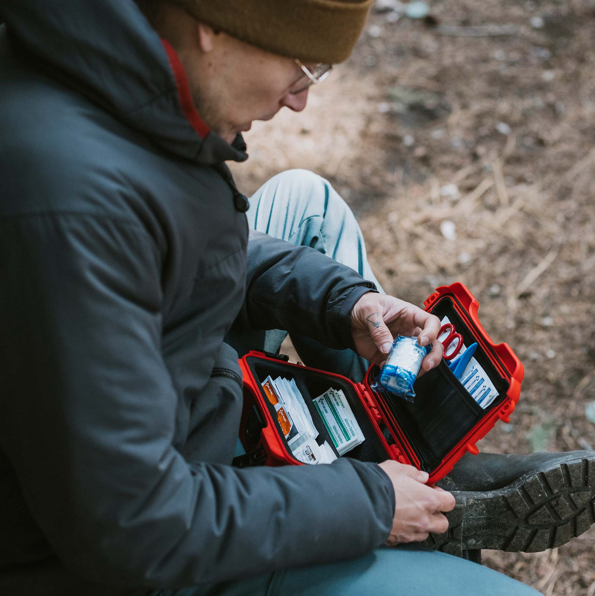 A person opens a red NANUK First Aid Case while seated on a rocky trail, surrounded by wilderness.