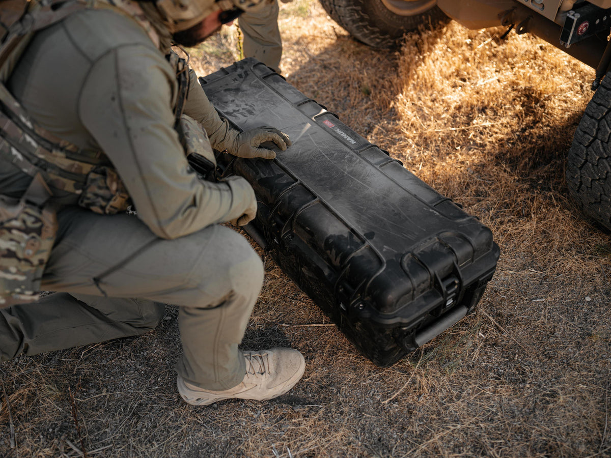 A black hard protective case lies on dry grass and dirt while a person in tactical gear kneels beside it outdoors near vehicle tires.