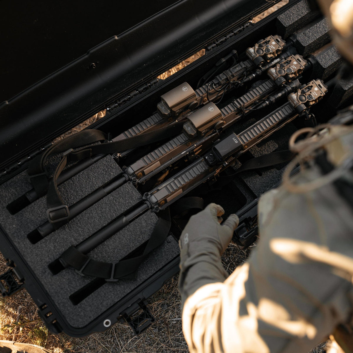 A black hard carrying case opened on the ground, showing three black tactical rifles with mounted optic accessories secured in foam inserts. A person wearing camouflage clothing and gloves is reaching into the case.