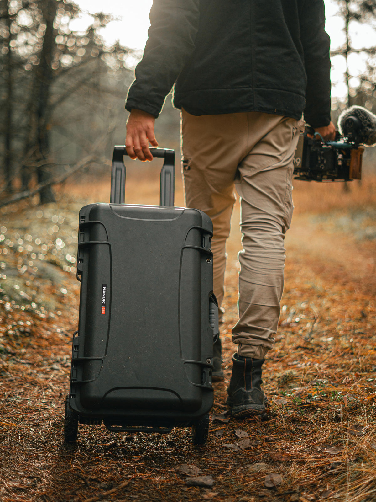 A person walking on a forest path pulling a large black rolling camera case with an extended handle, while holding a camera in the other hand.
