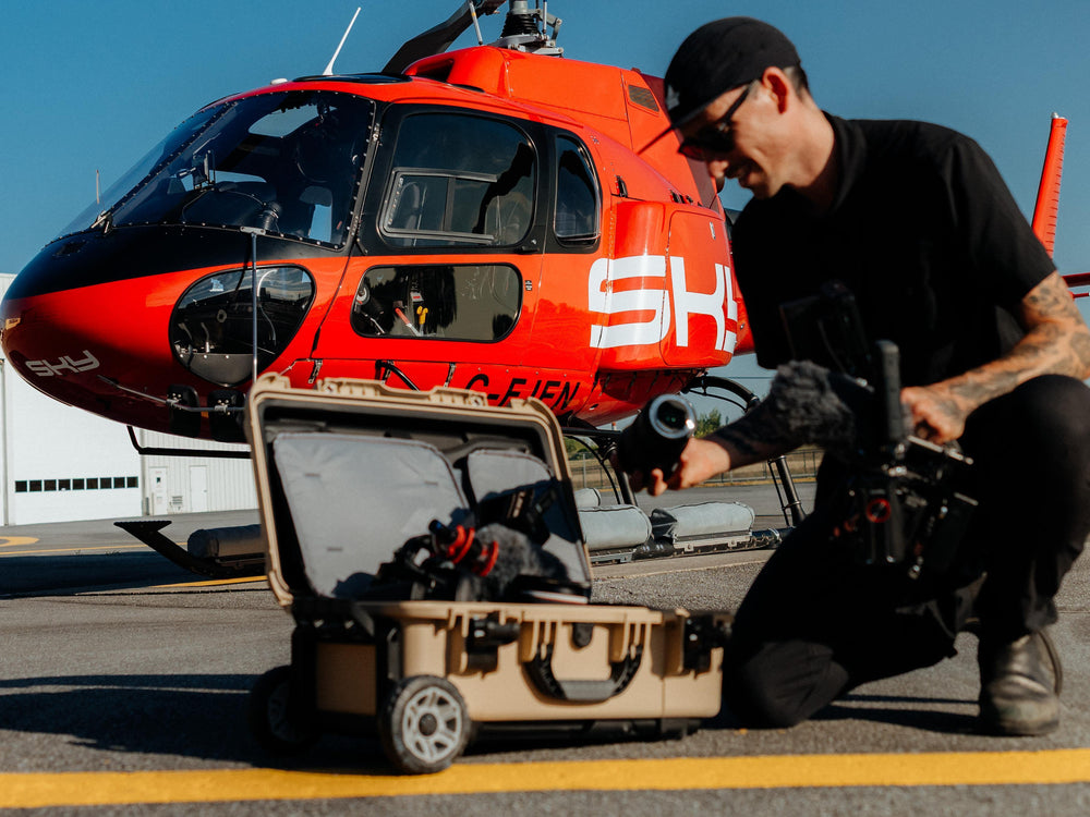 A man in black clothing kneels on an airport tarmac in front of a bright red helicopter, holding a camera controller next to an open beige hard-shell suitcase containing a drone.