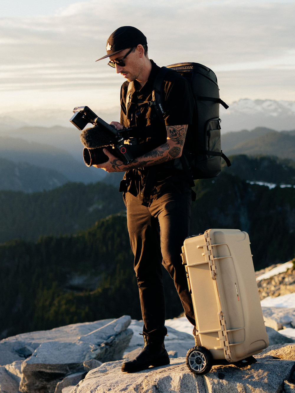 A man standing on a mountain peak with a camera and a large black backpack, next to a beige hard-shell rolling suitcase. The background shows a scenic mountain landscape during golden hour.