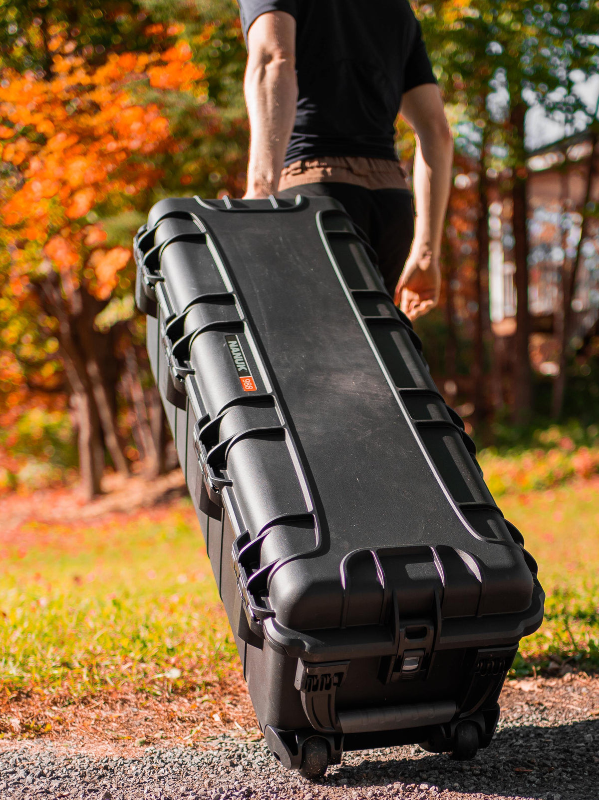 A person outdoors in an autumn park setting pulls a large black hard-sided rolling case with wheels. The case has visible Zennit branding and latches.