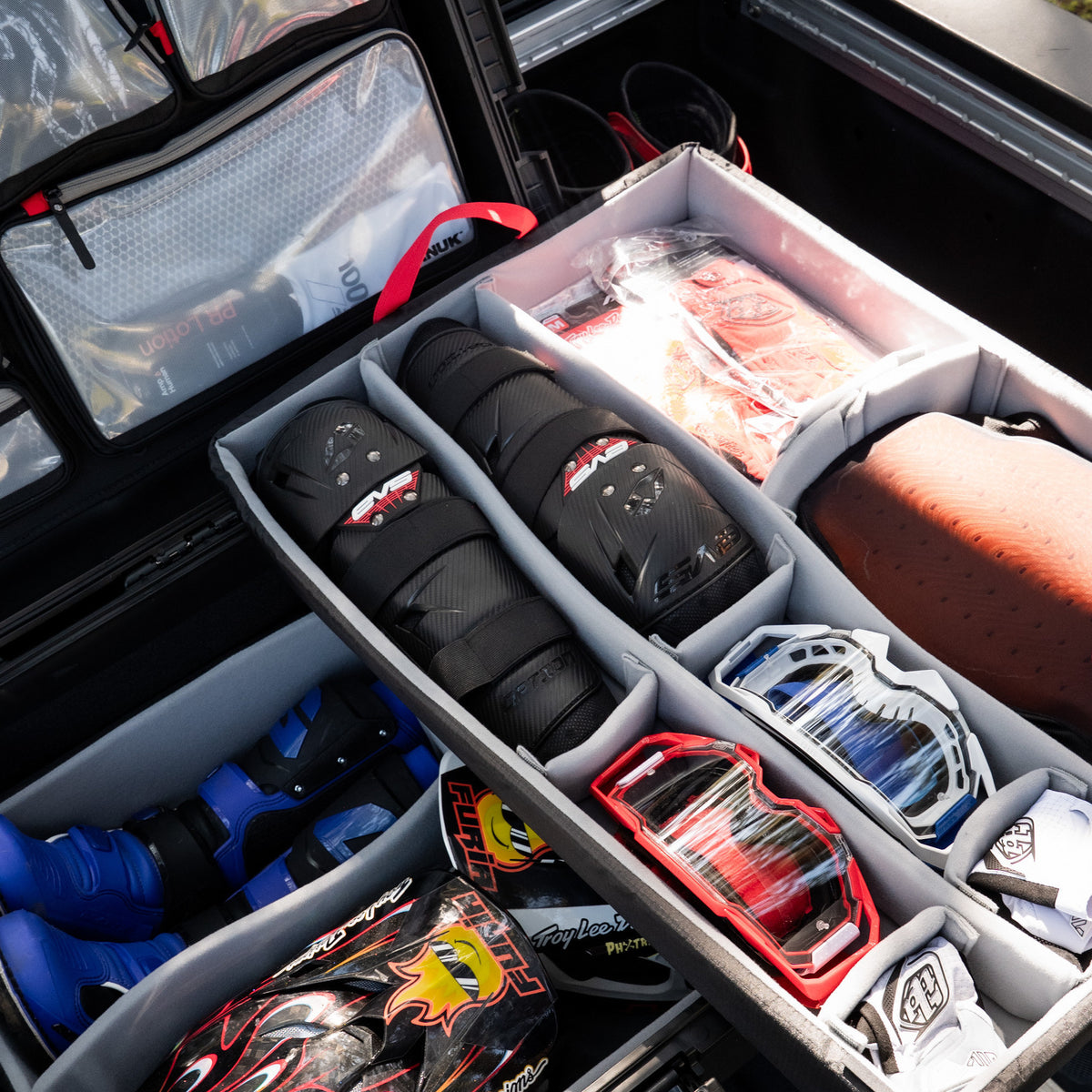 A close-up of a gear organizer tray inside a vehicle or case, filled with protective sports equipment including black knee pads, blue boots, red and blue goggles, and other helmets or accessories.