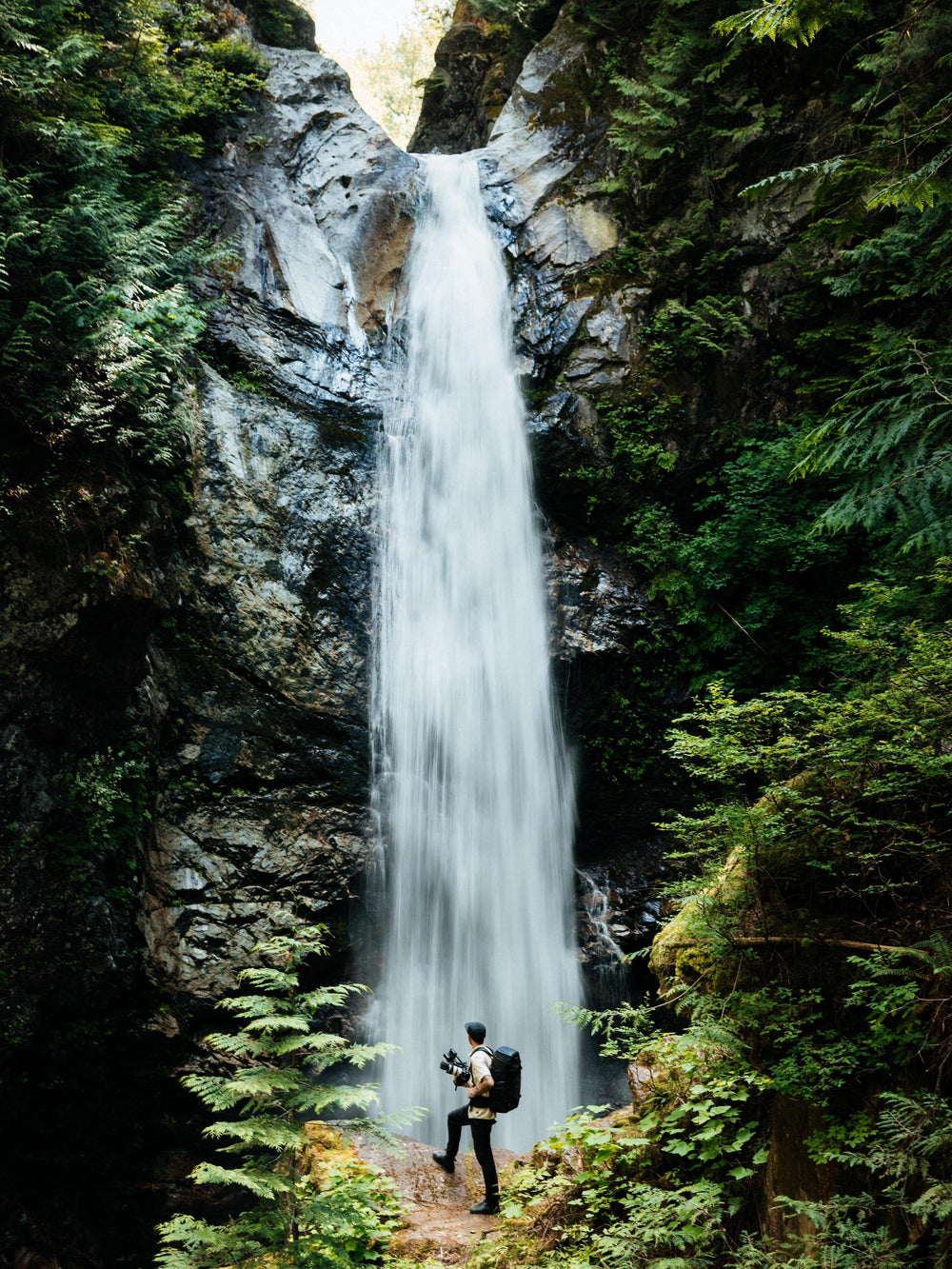 A tall waterfall cascades down a rocky cliff surrounded by dense green forest, with a small person standing near the base holding a camera.
