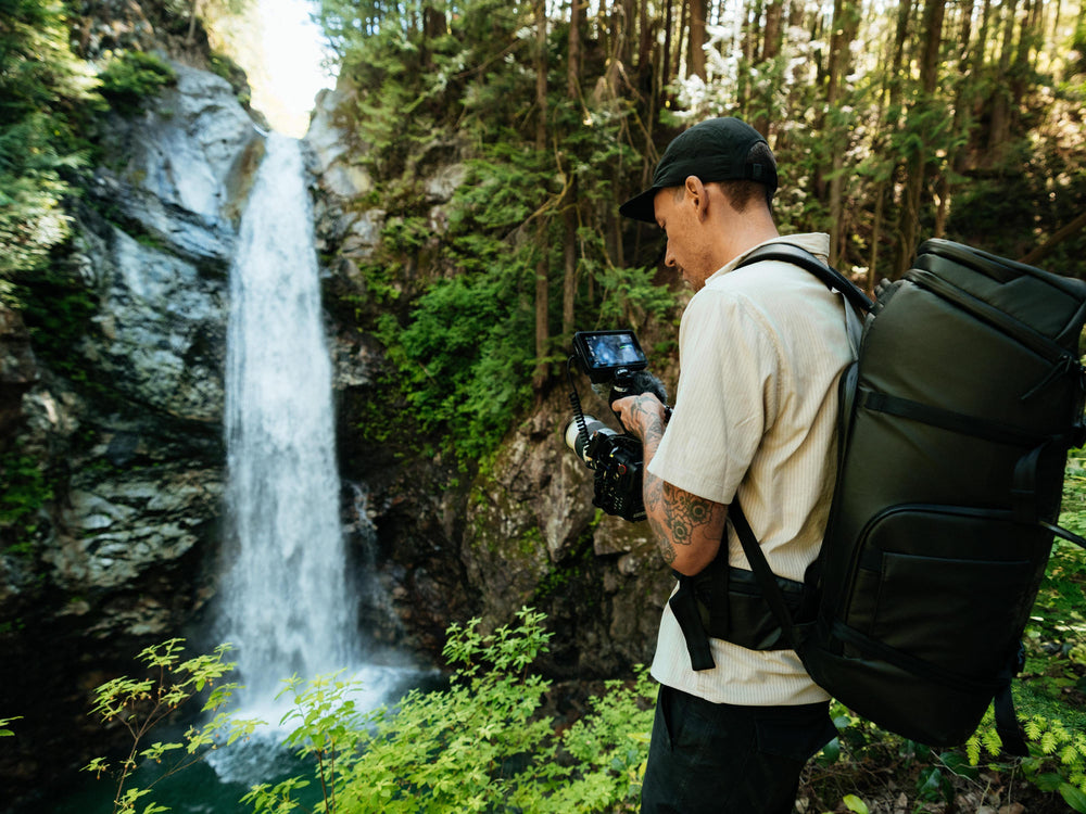 A man wearing a beige t-shirt, black cap, and a large black backpack stands in a lush forest next to a waterfall, holding a camera with a screen attached.