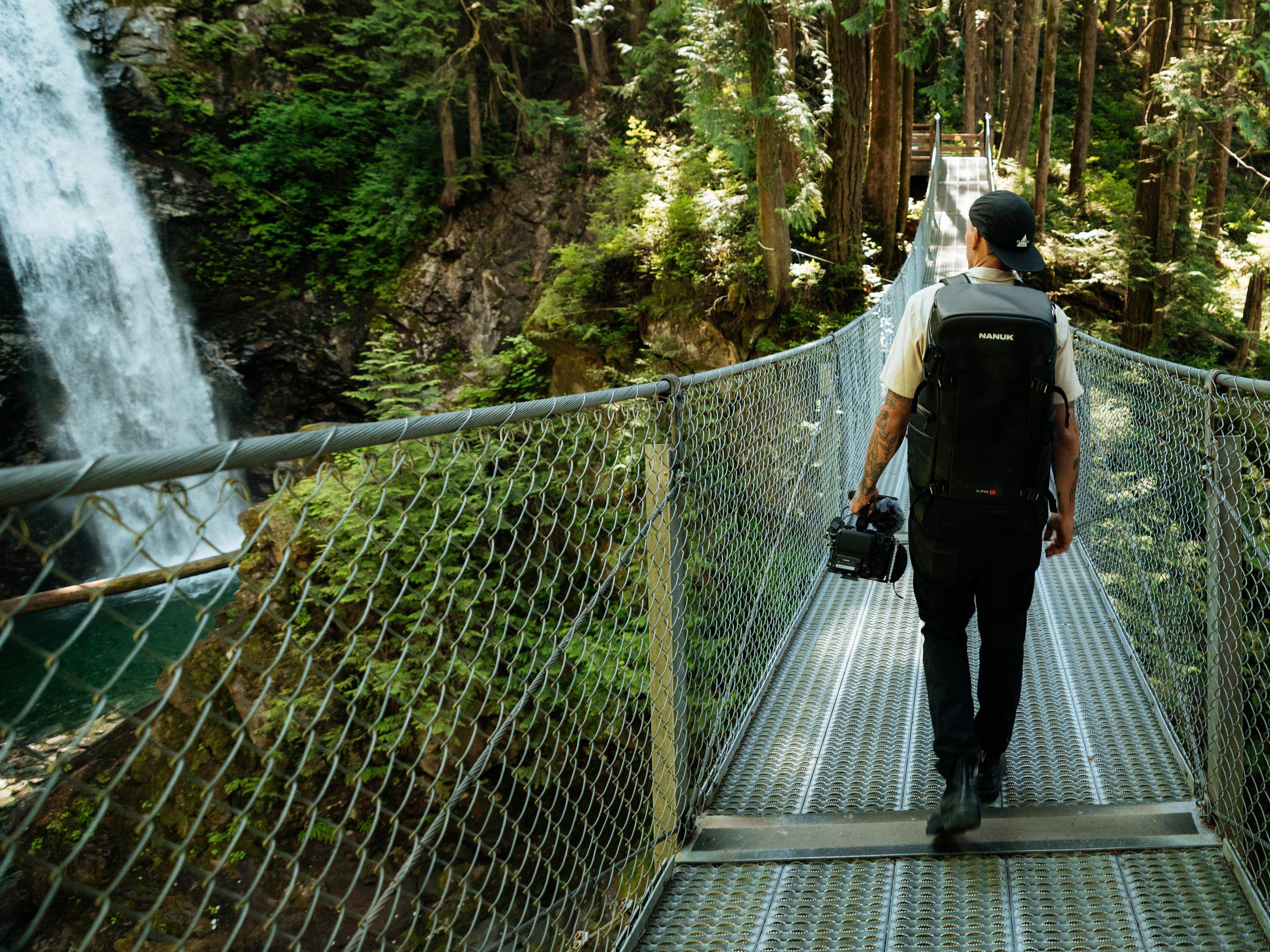 A man wearing a black backpack and walking across a suspension bridge in a forest, with a waterfall visible on the left side. He is carrying a camera.