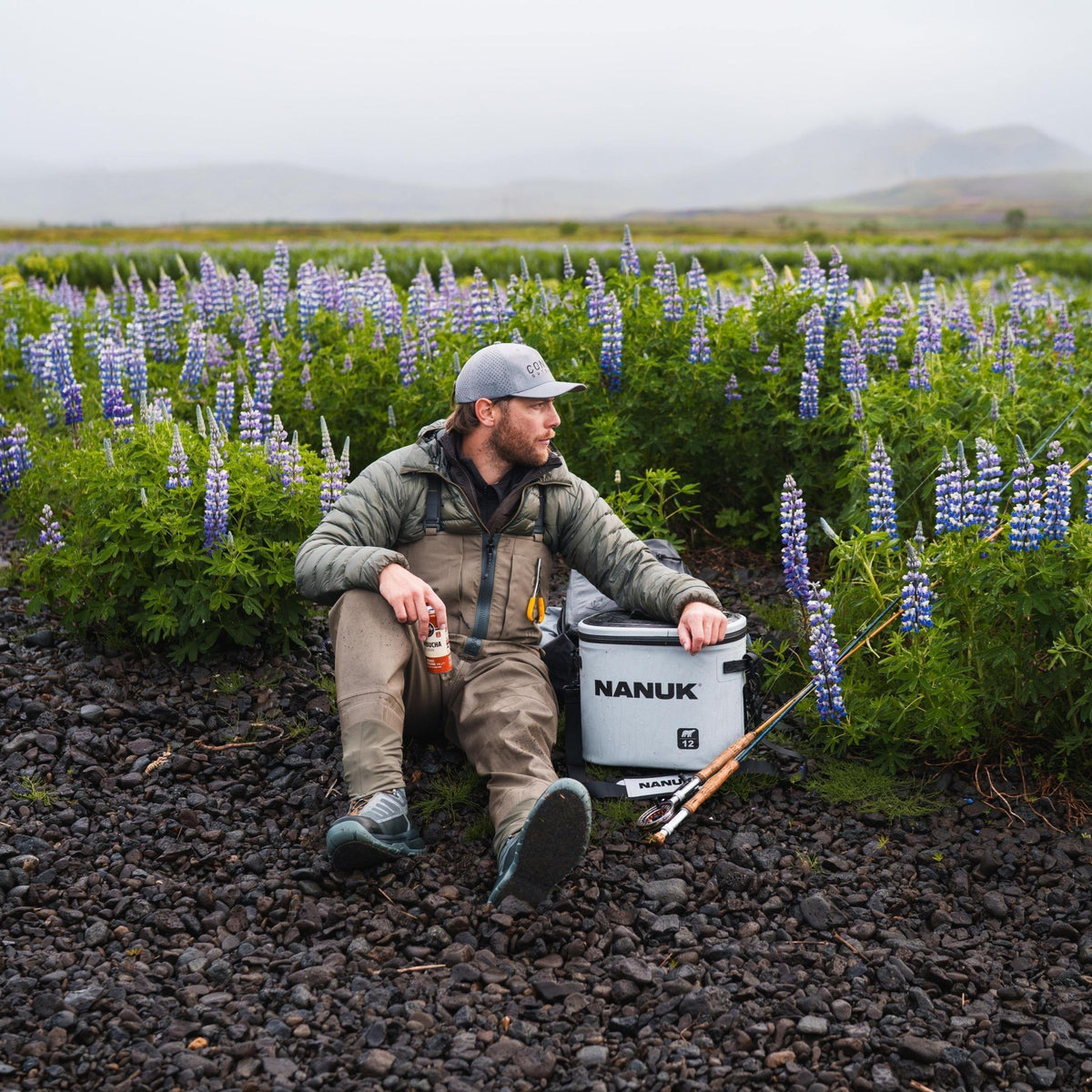 A man in outdoor fishing attire sits on dark gravel beside a white NANUK cooler, with a fishing rod and a field of purple lupines in the background.