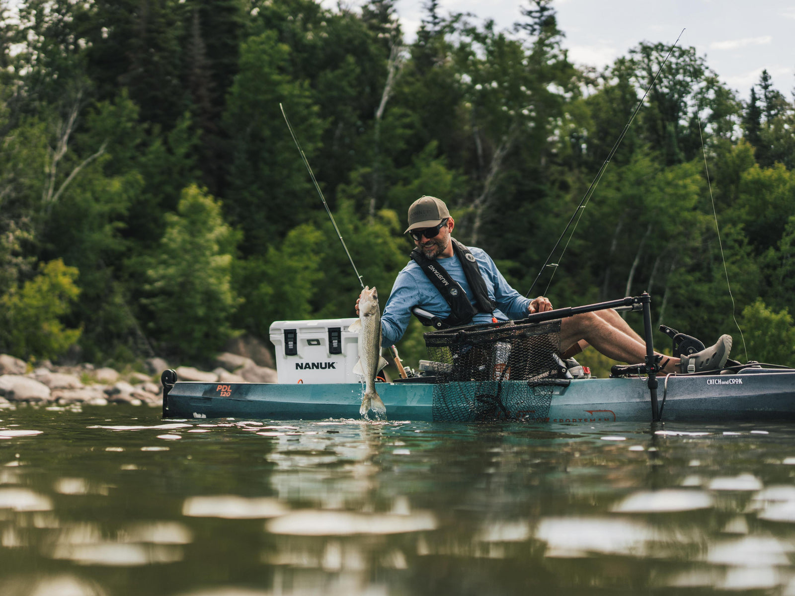 A man fishing from a teal Sitka kayak on a river, holding a fish near a white Nanuk cooler, with a forest background.