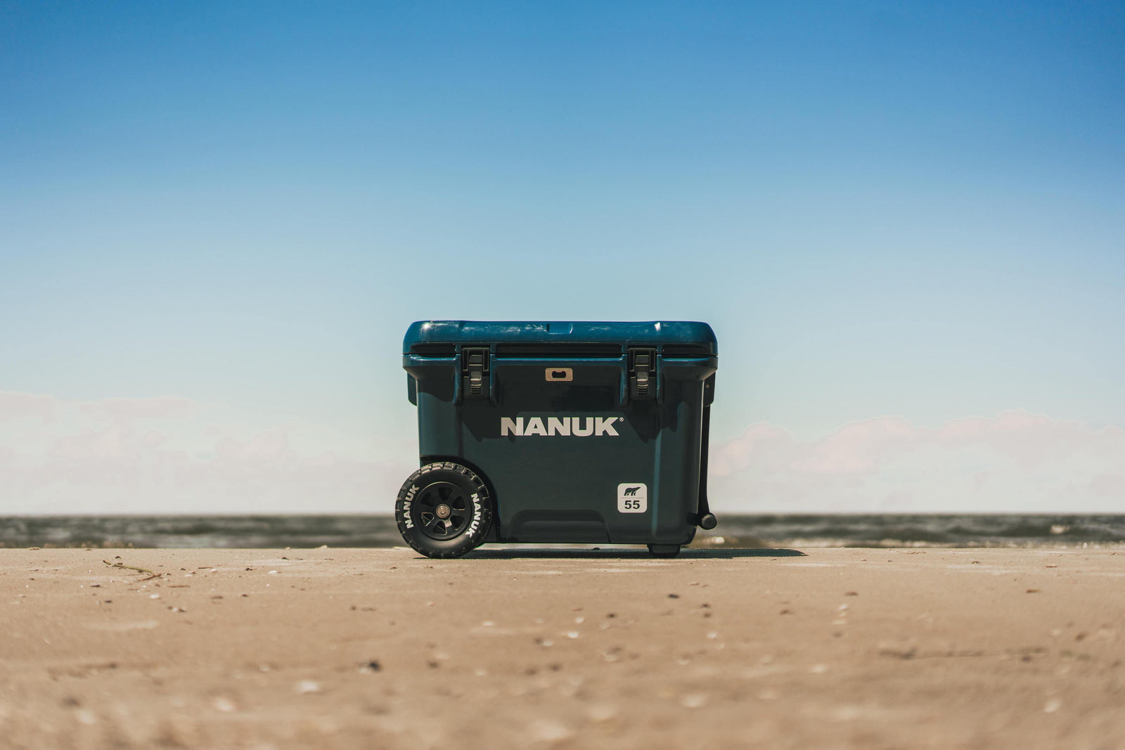 A dark teal Nanuk cooler with wheels, standing on a sandy beach under a clear blue sky.