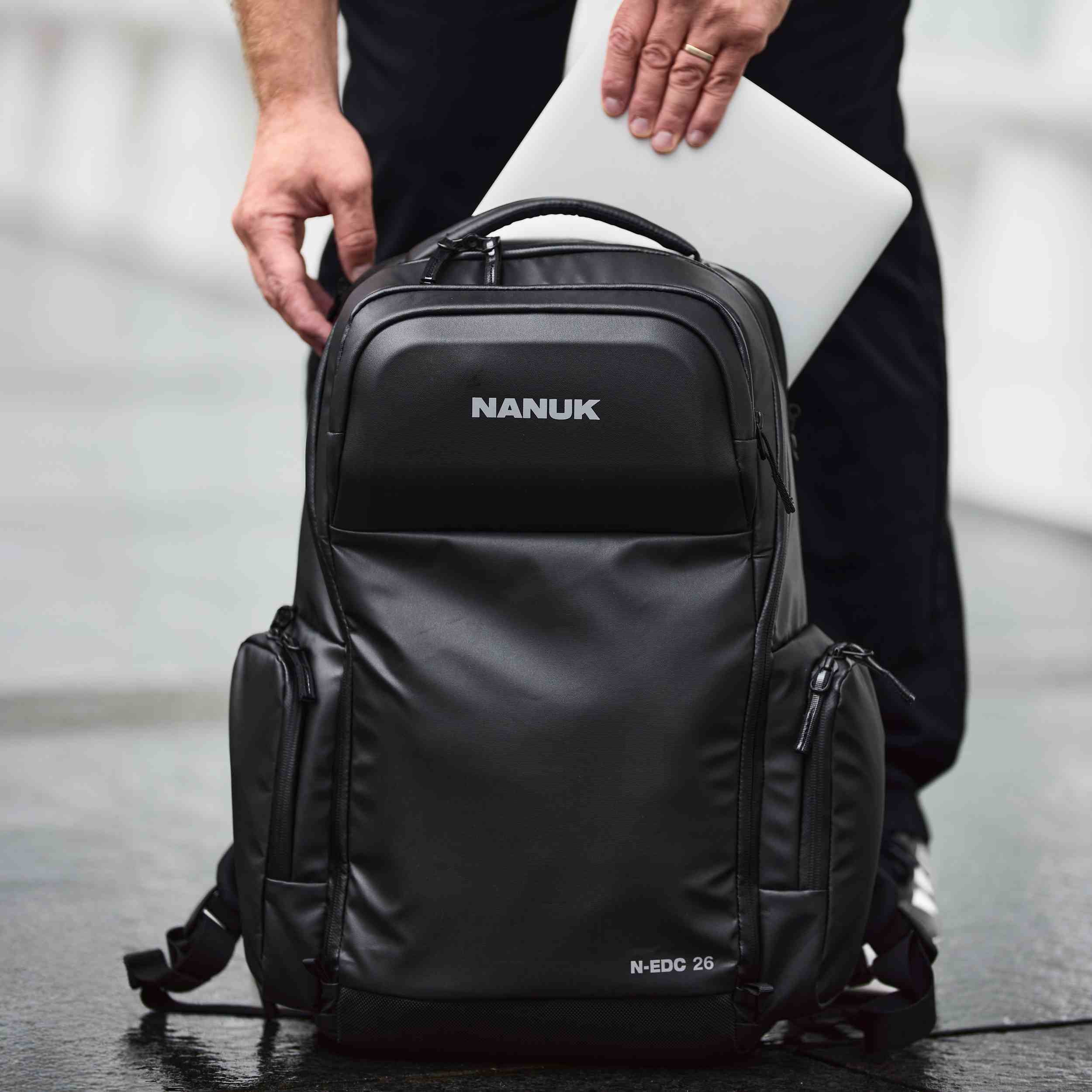 Black Nanuk backpack with a person holding a laptop inside, on a blurred background.