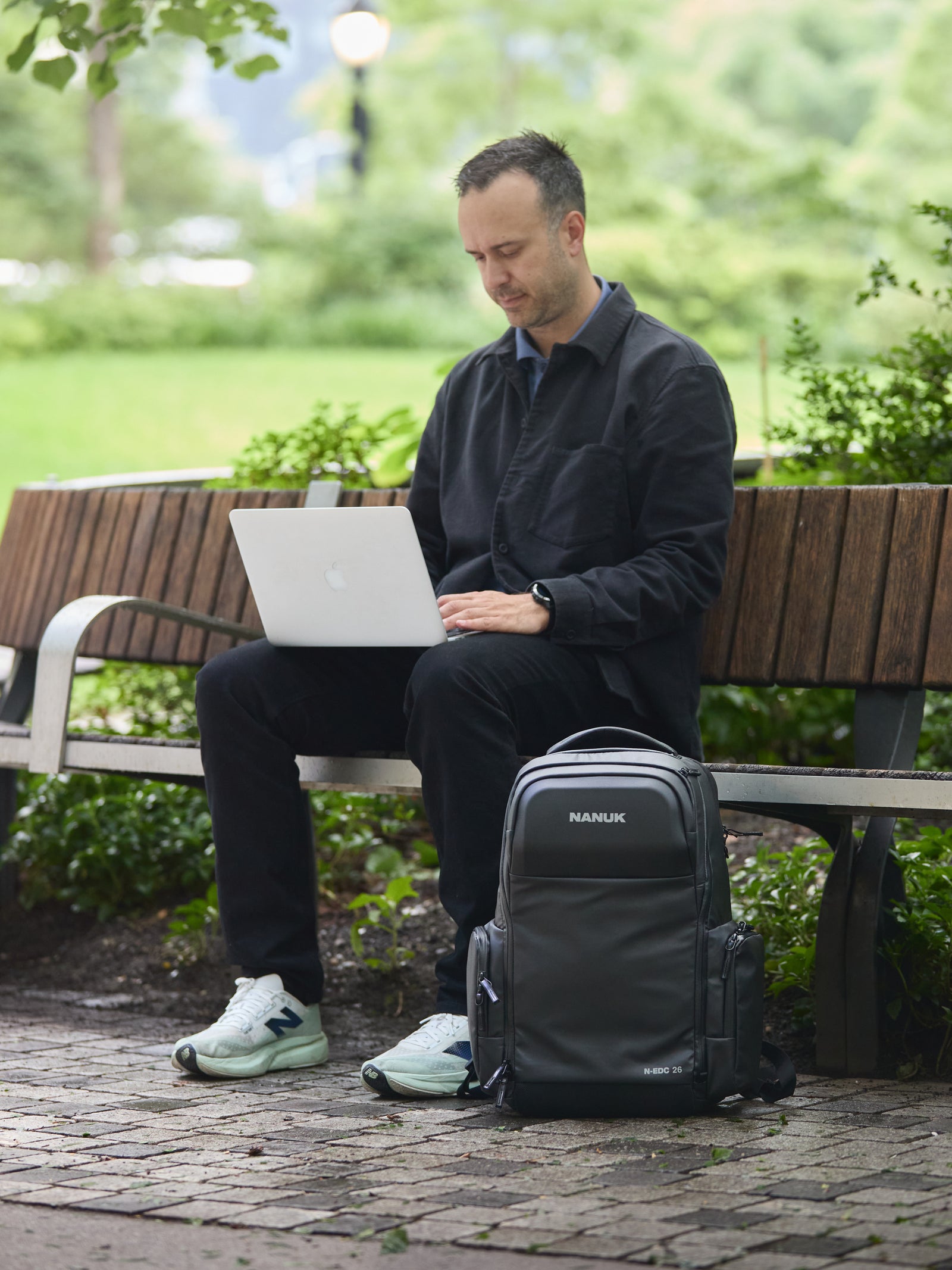 Man sitting on a bench in a park using a laptop with a Naukri backpack next to him.