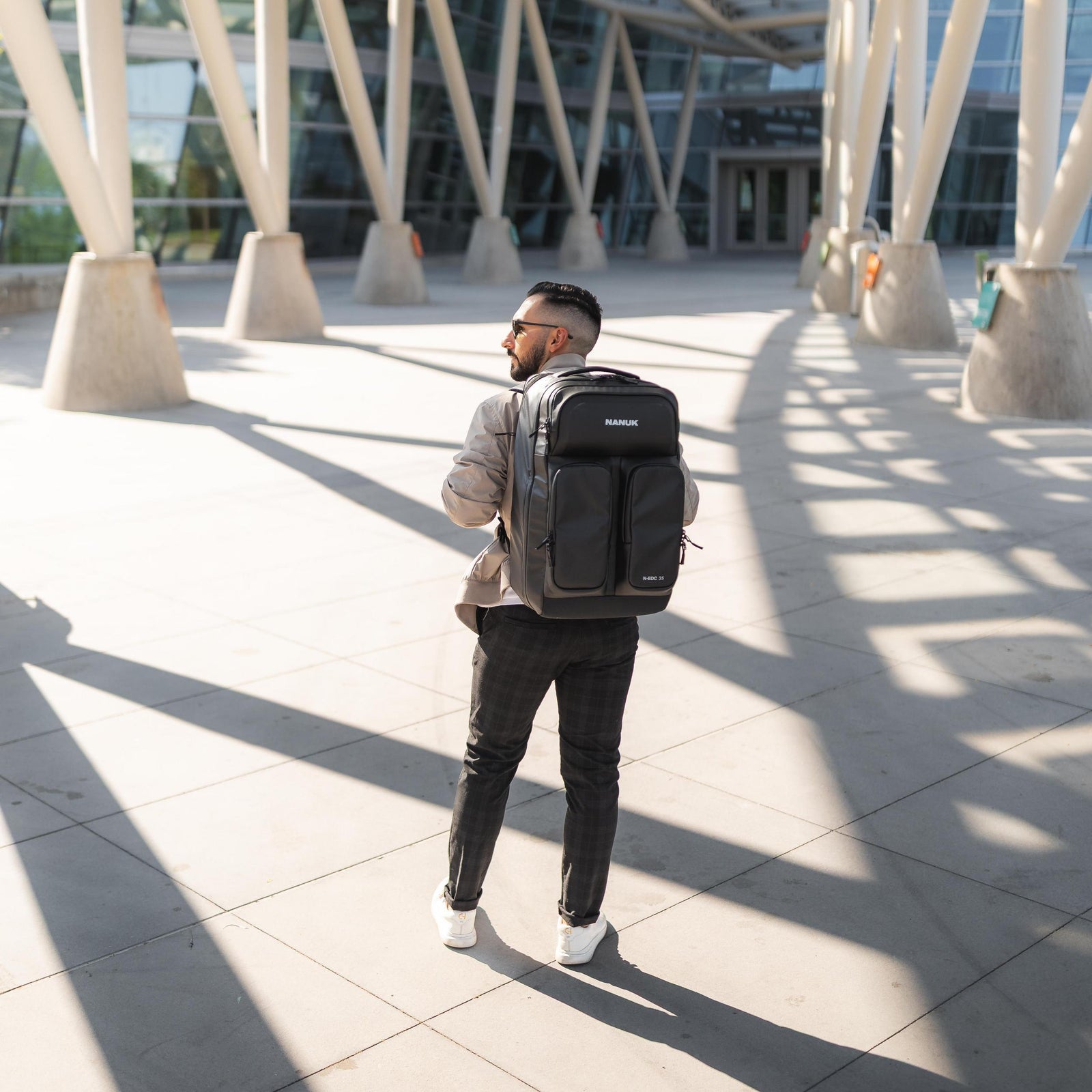 Man with a backpack walking on a sunlit pavement with modern architecture in the background
