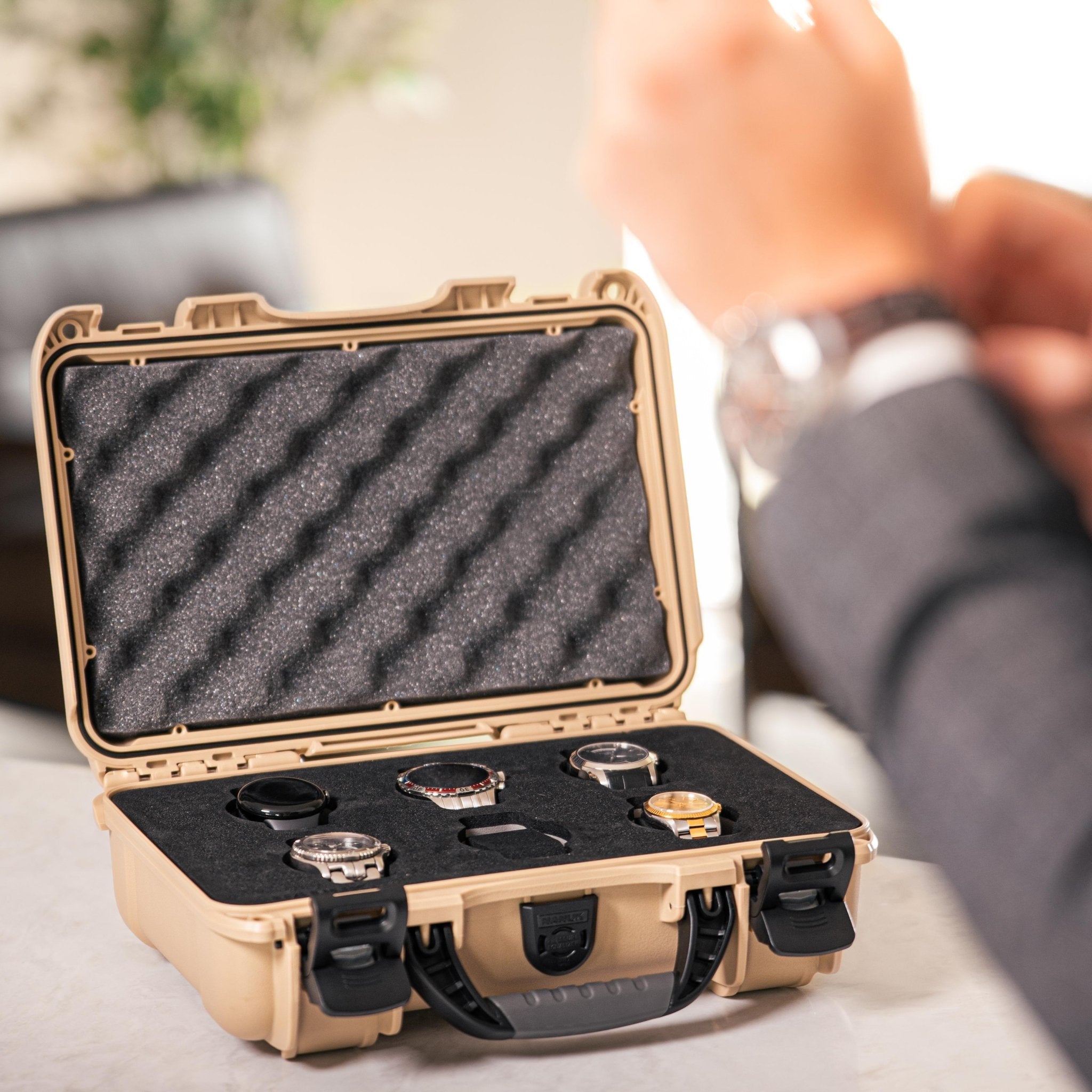 A tan hard case with black foam interior holding several wristwatches, shown open in a real indoor setting with a person's arm blurred in the background.