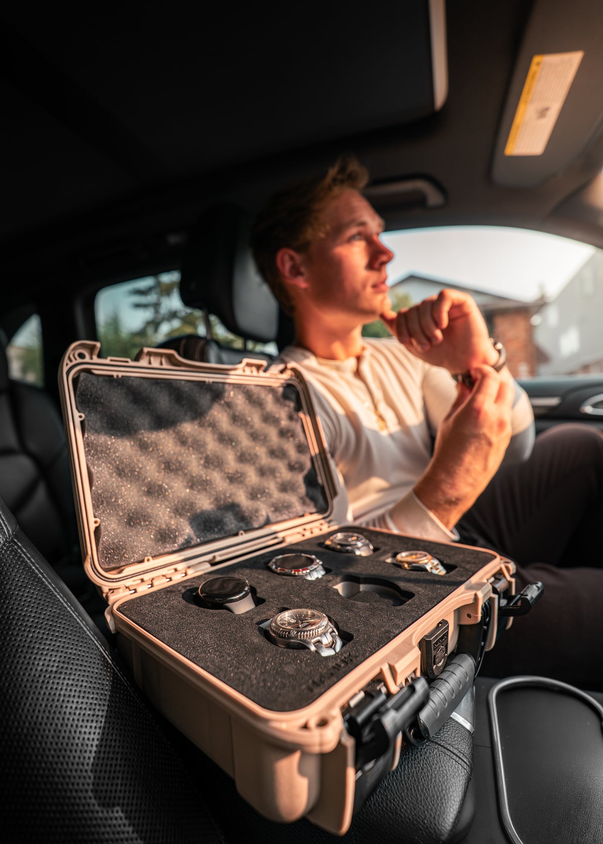 A man sitting inside a car looks at an open hard case on the seat, which contains several watches arranged in a foam insert. The case is tan with a black foam interior.