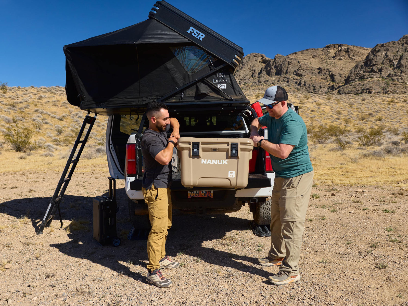 Two men setting up a rooftop tent on a truck in a desert landscape.