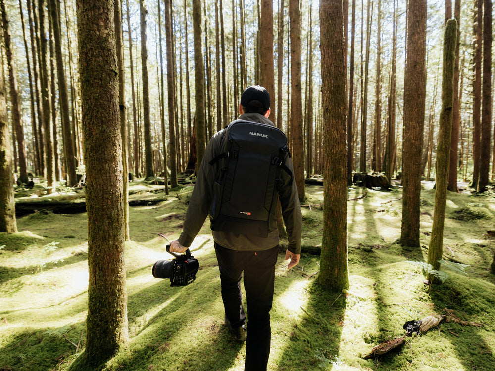 Person walking through a forest with a backpack and camera, surrounded by tall trees.